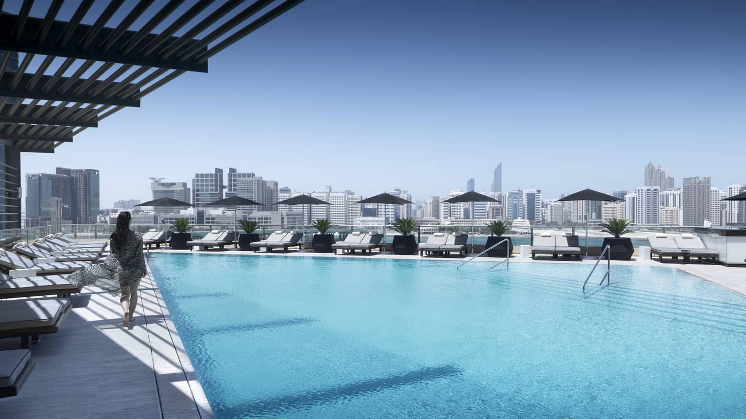 Back view of woman walking past lounge chairs on deck past blue swimming pool on sunny rooftop patio