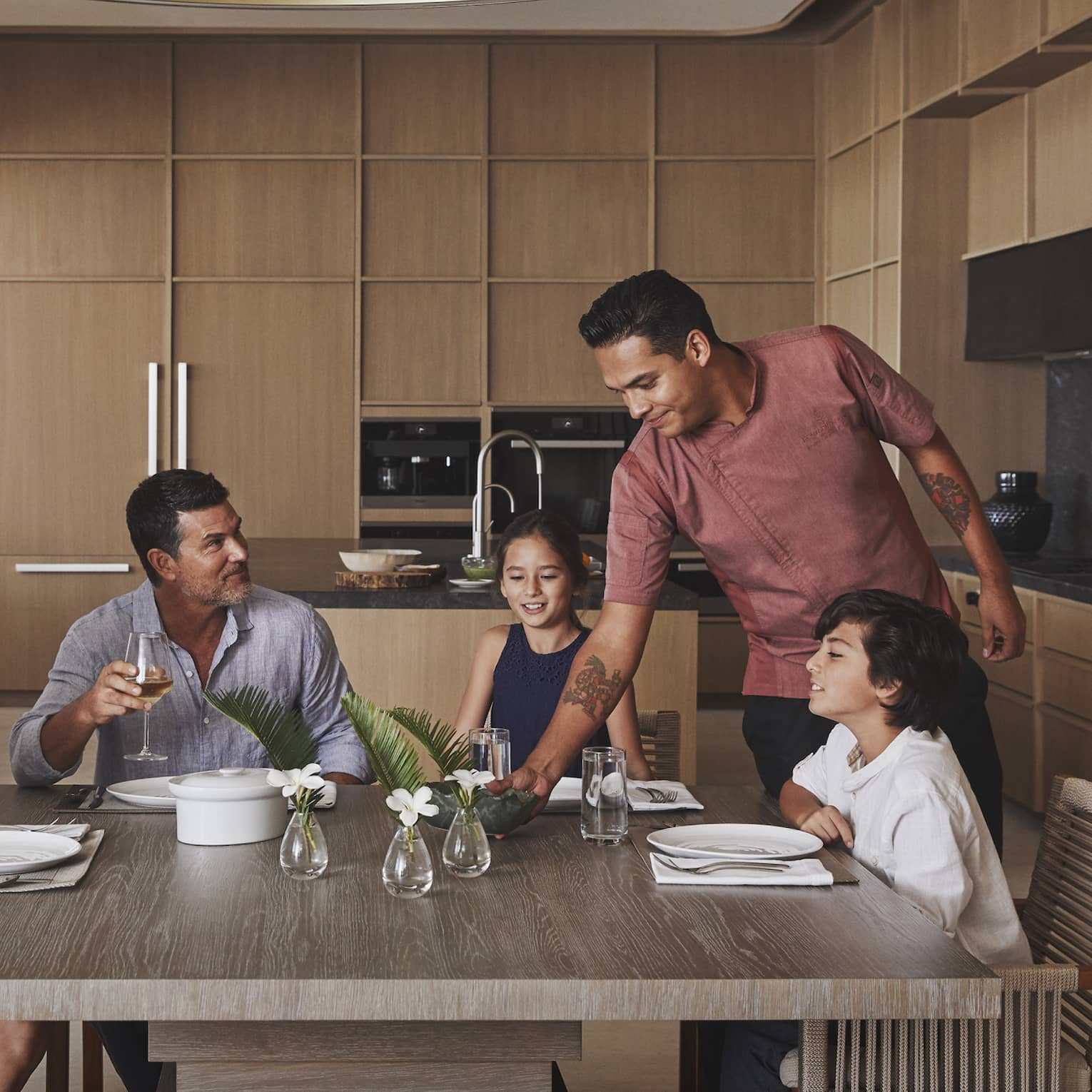 Four guests sitting at the table in a villa while a chef places a dish on the table.