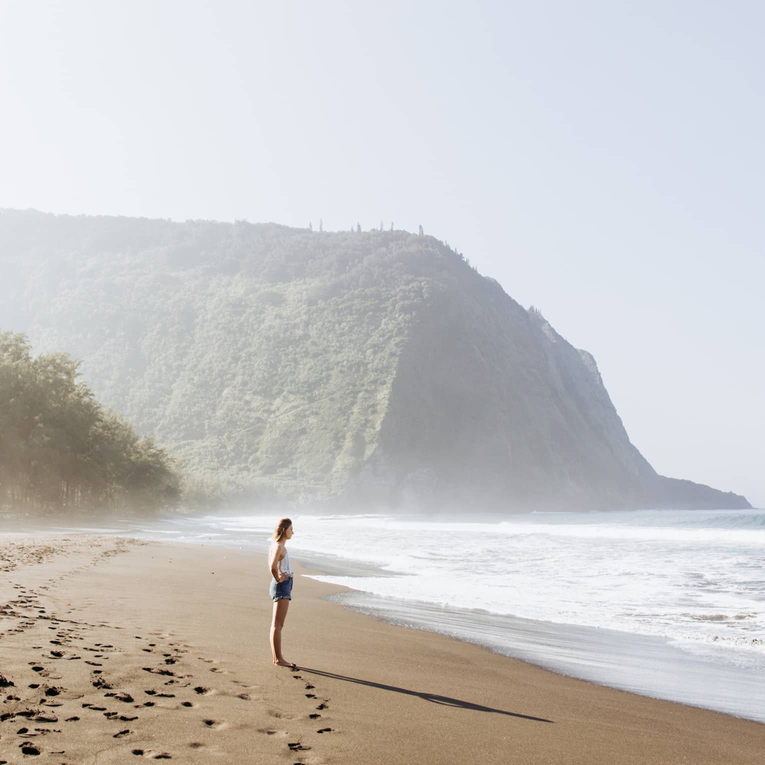 On caramel-coloured sand dotted by footprints, a beachgoer gazes into the ocean; a lush, misty mountain in the background.