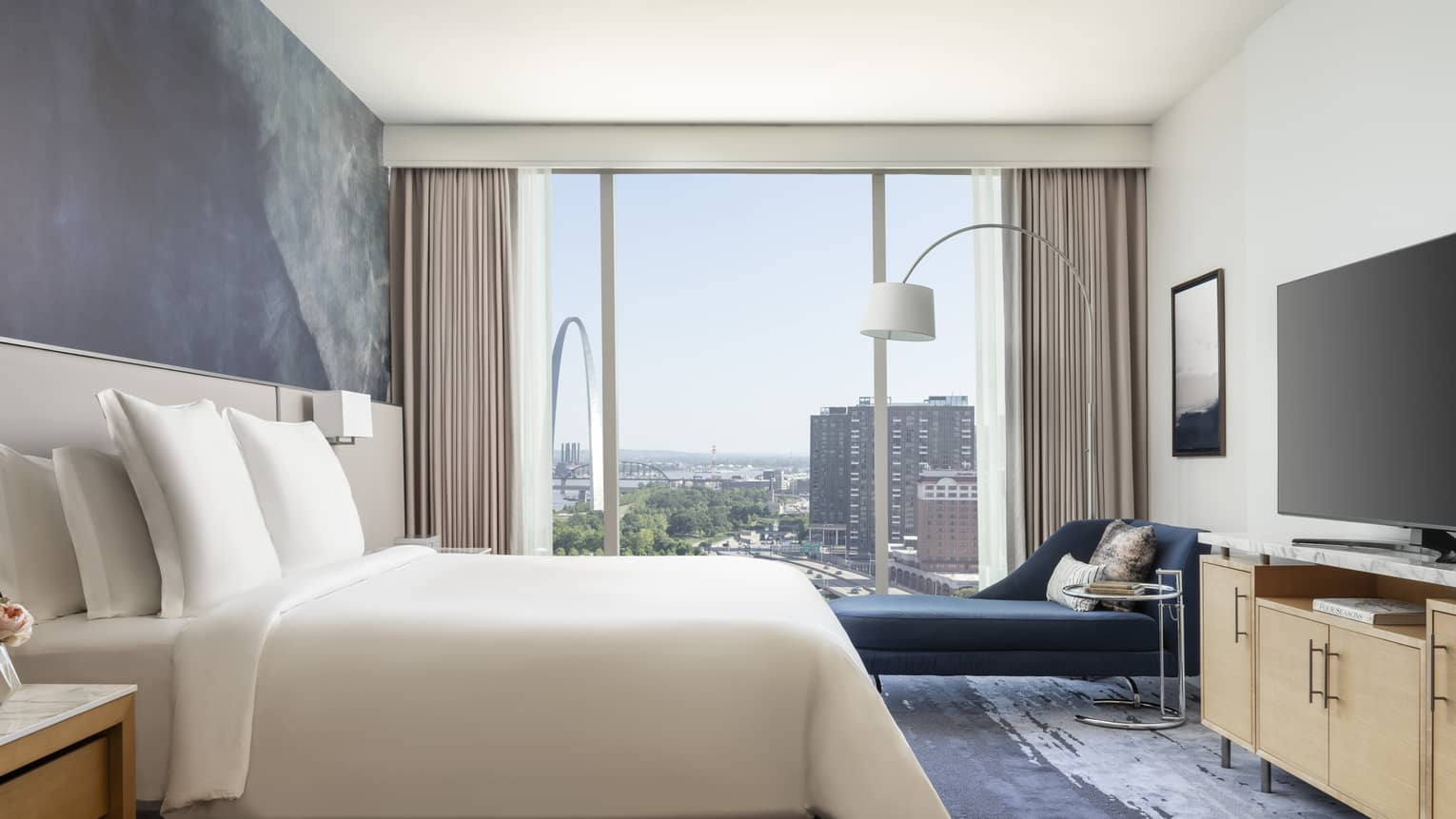 Hotel room with king bed, dresser, TV, dark blue chaise next to wall of windows