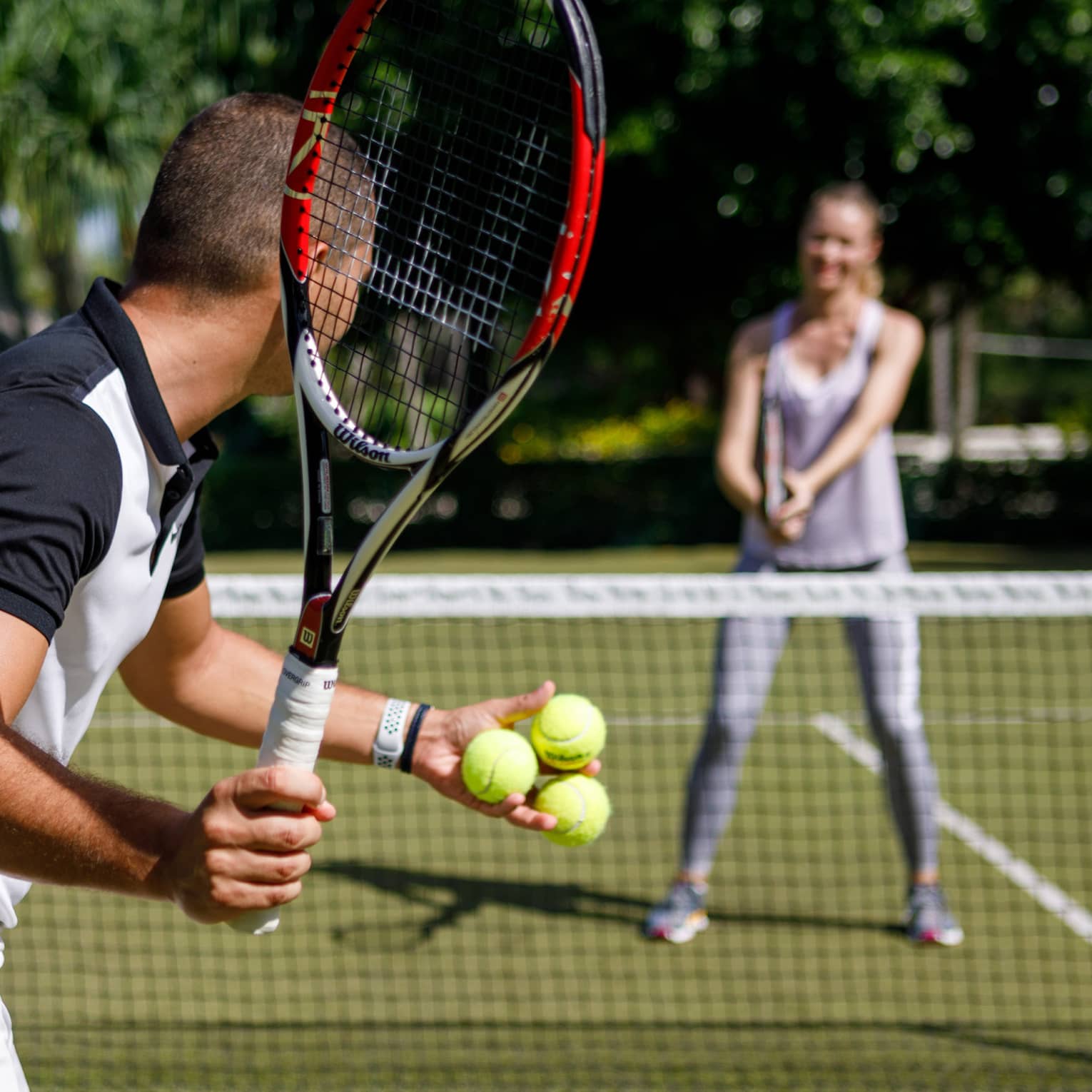 Couple playing tennis on court, man prepares to serve tennis balls to woman ready to swing