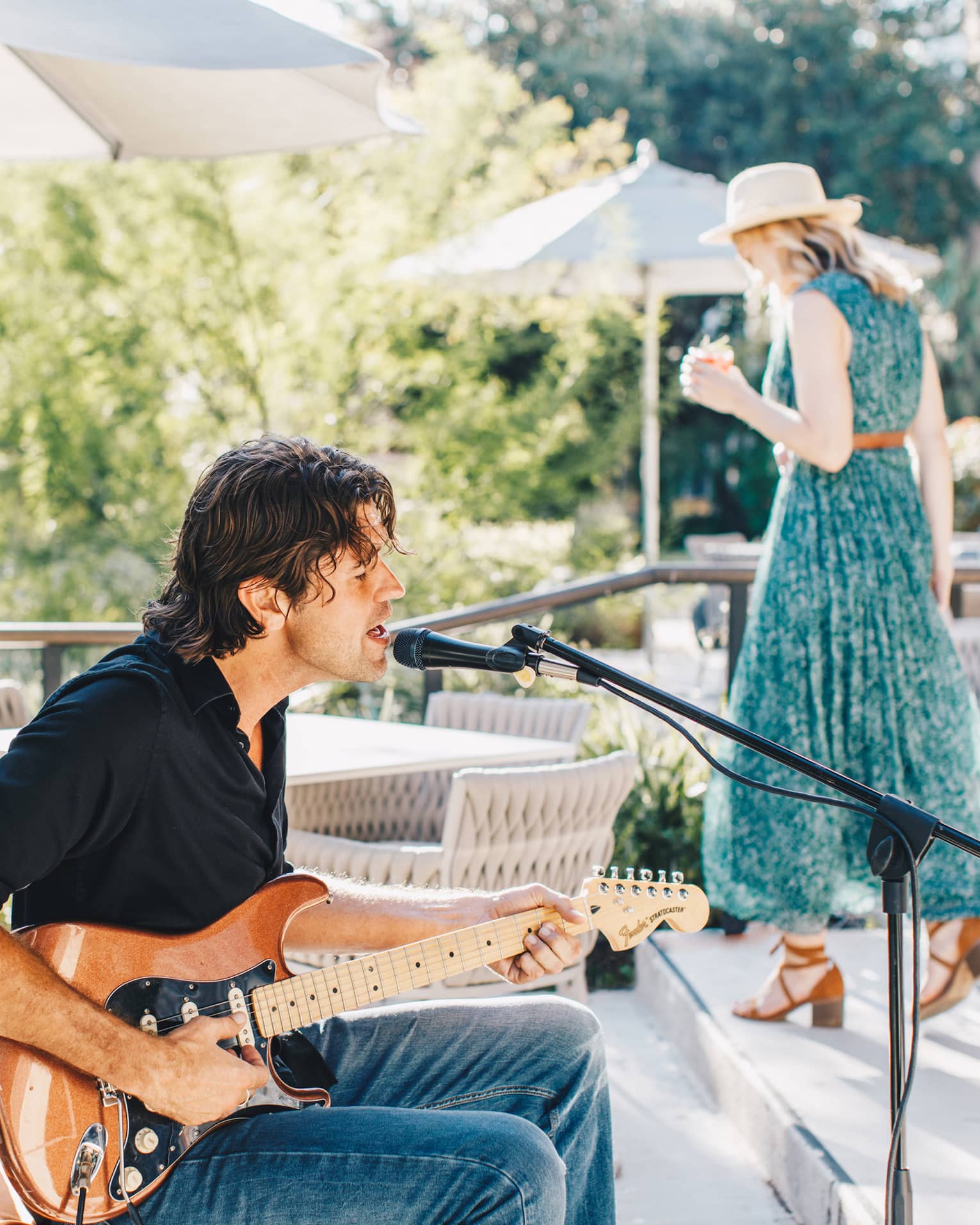A man sitting and playing guitar while a woman in a green dress walks behind him.