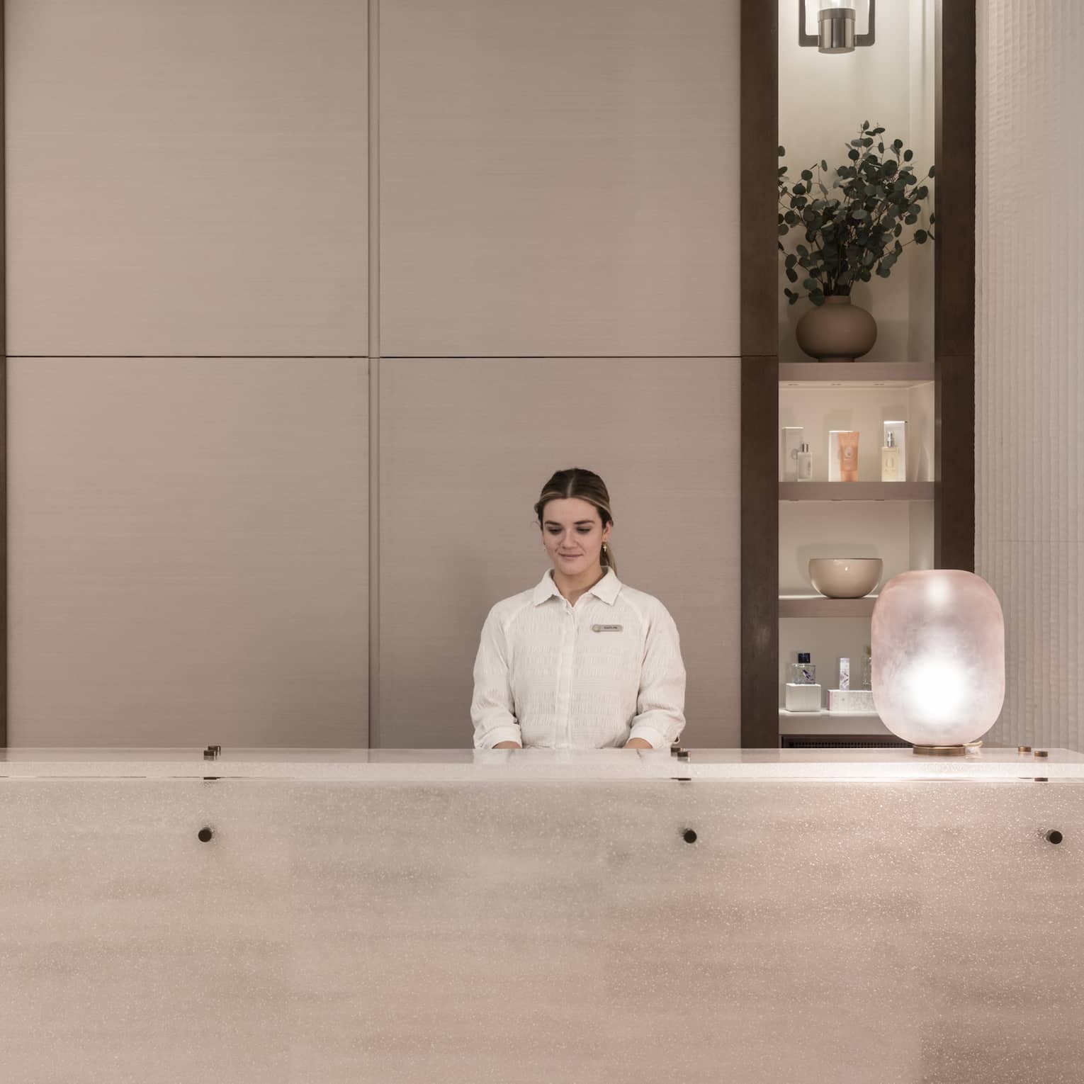 Receptionist in white shirt stands behind white marble spa reception desk with shelving display in backdrop
