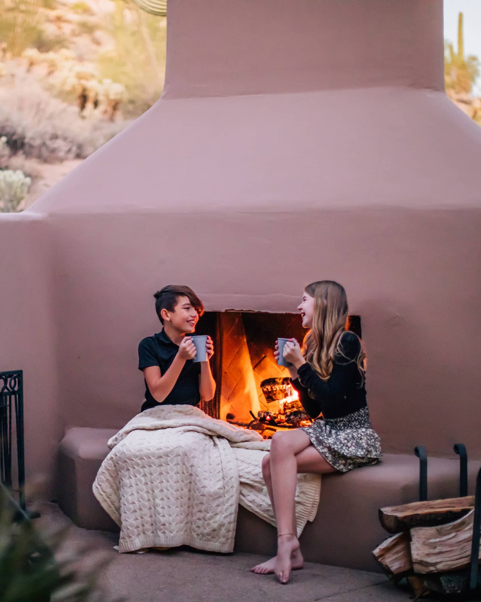 Two people sitting by a fire outside while drinking from mugs.