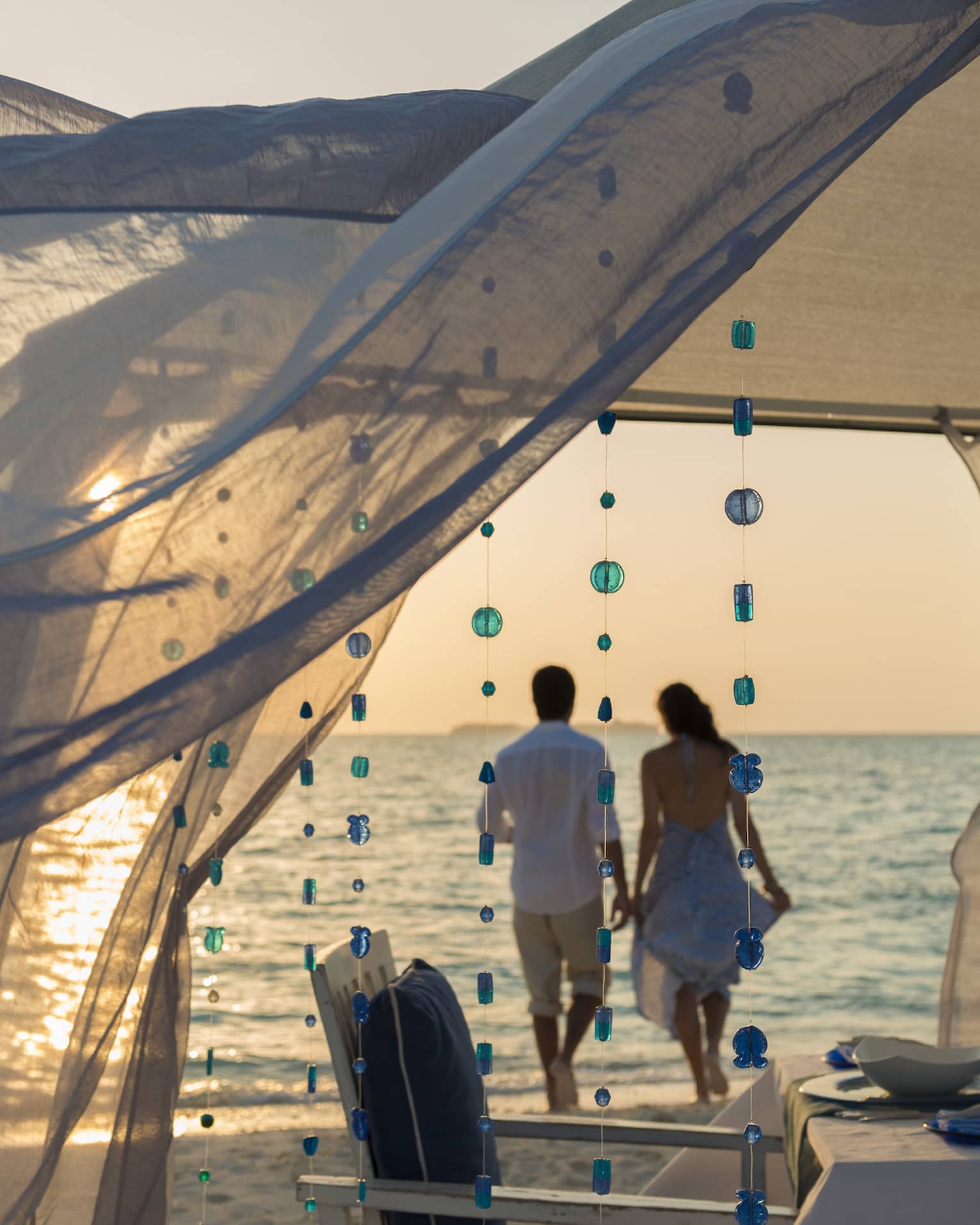 Sheer curtains blow in breeze, small blue beads hang from string, woman and man hold hands on beach in background