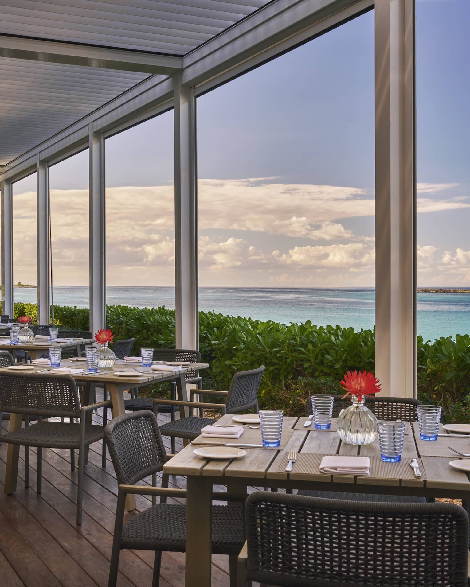 An outdoor eating area with square wood tables next to plants and the ocean.