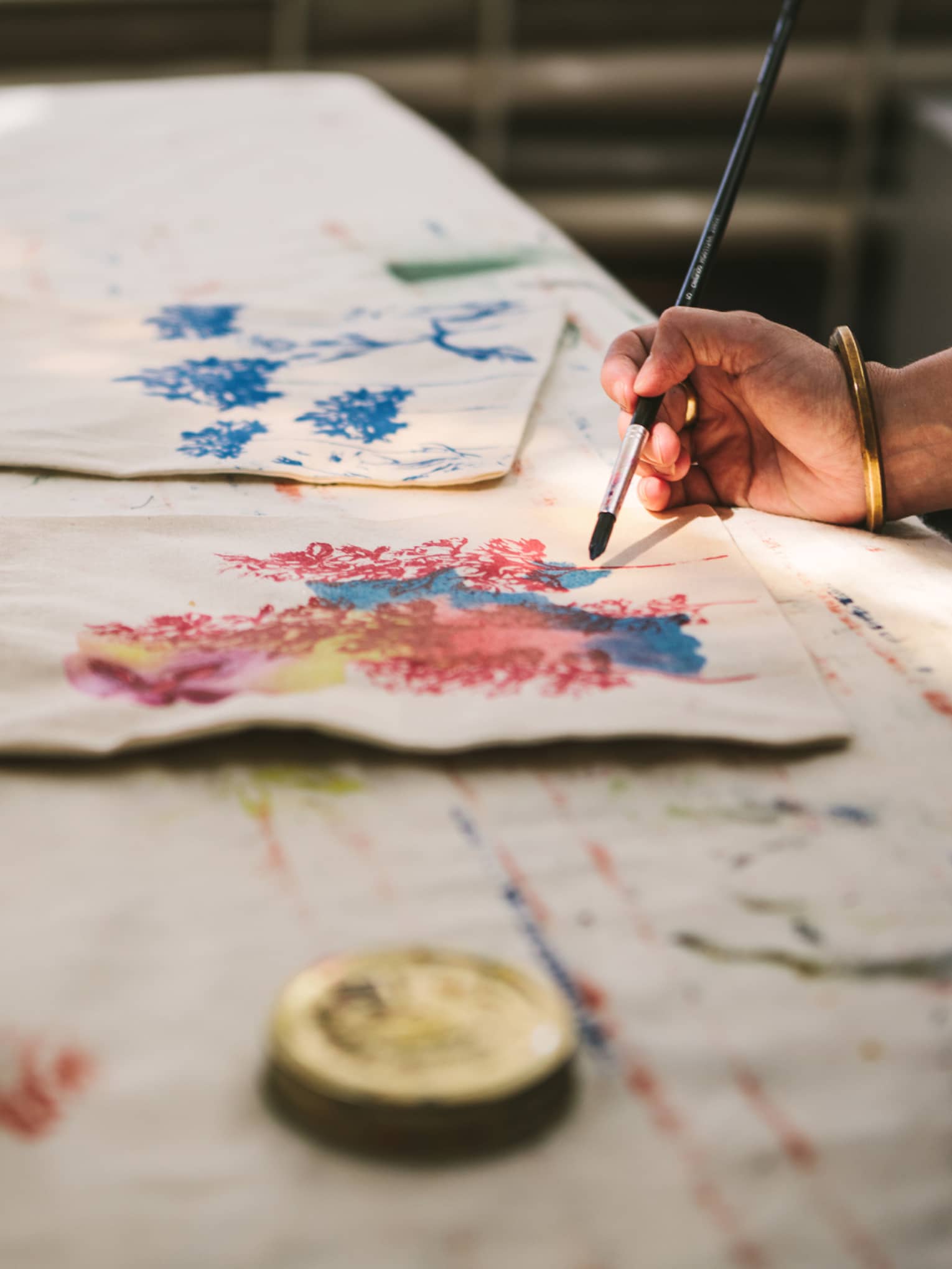 Close-up of a gold-bangled hand wielding a long brush to paint an intricate pattern on a cloth tote bag.