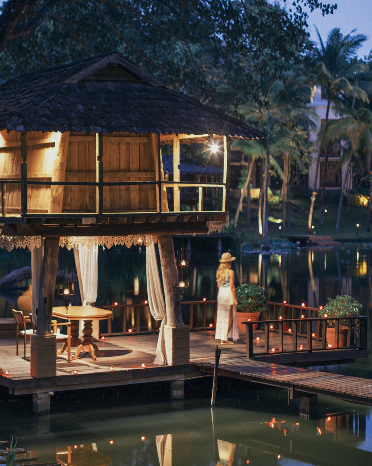 A guest looks out at a serene river at dusk, on the platform of a wooden hut, as lanterns and twinkling lights dot the scene.
