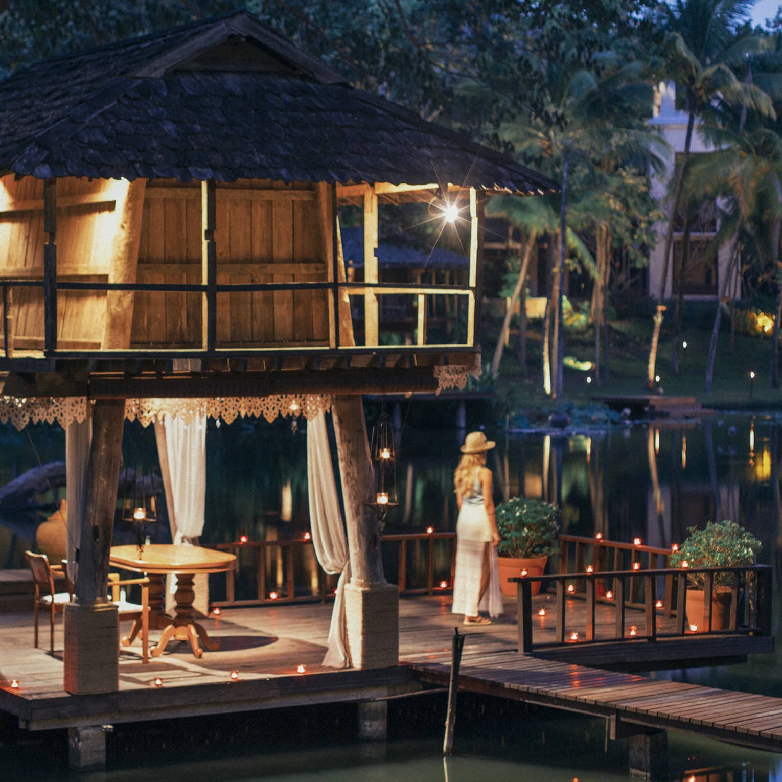 Person standing on a dock beside a candlelit lakeside pavilion with a thatched roof and draped curtains at dusk.