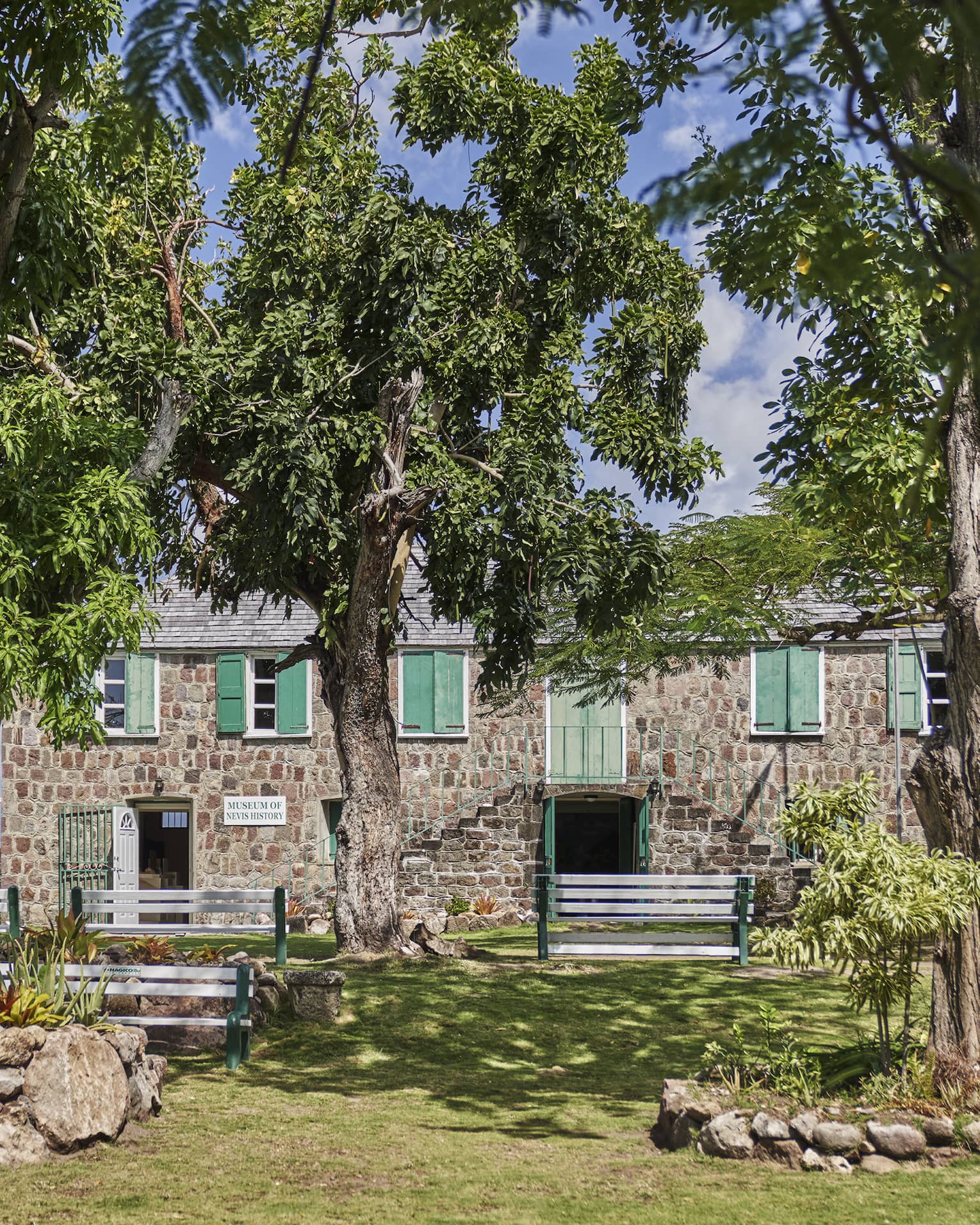 A two-storey stone building with green doors and shutters, seen across a lawn and behind trees, flags blowing behind it.