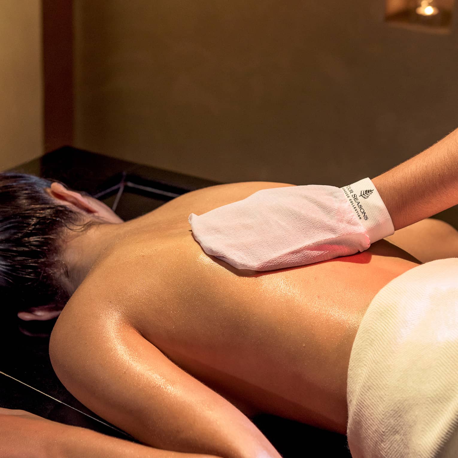 Woman lies face-down on spa table while hand with white mitten massages her back