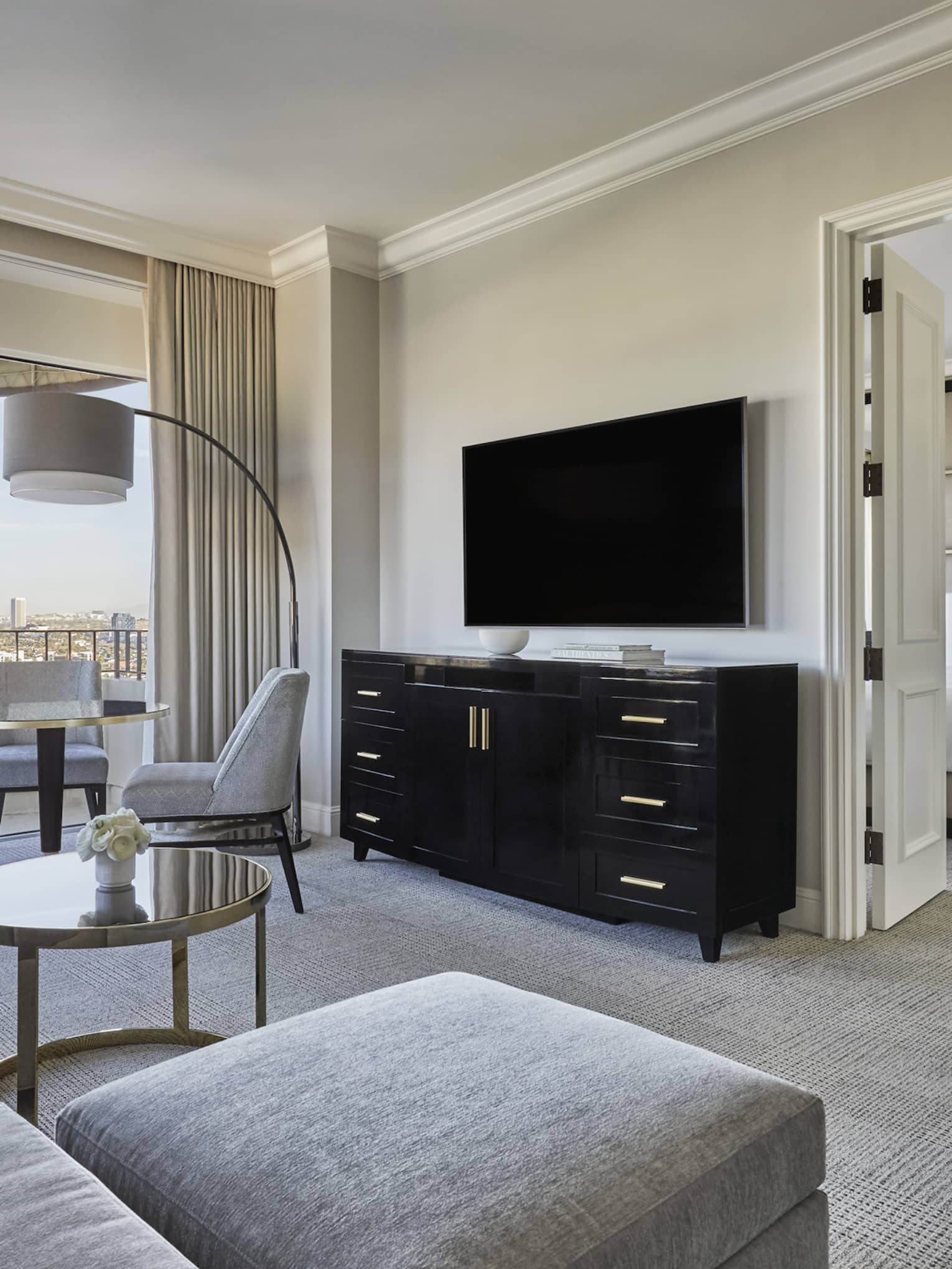 A hotel room with a grey couch, dresser, tv, lamp, small table, bed with white sheets, and large window.