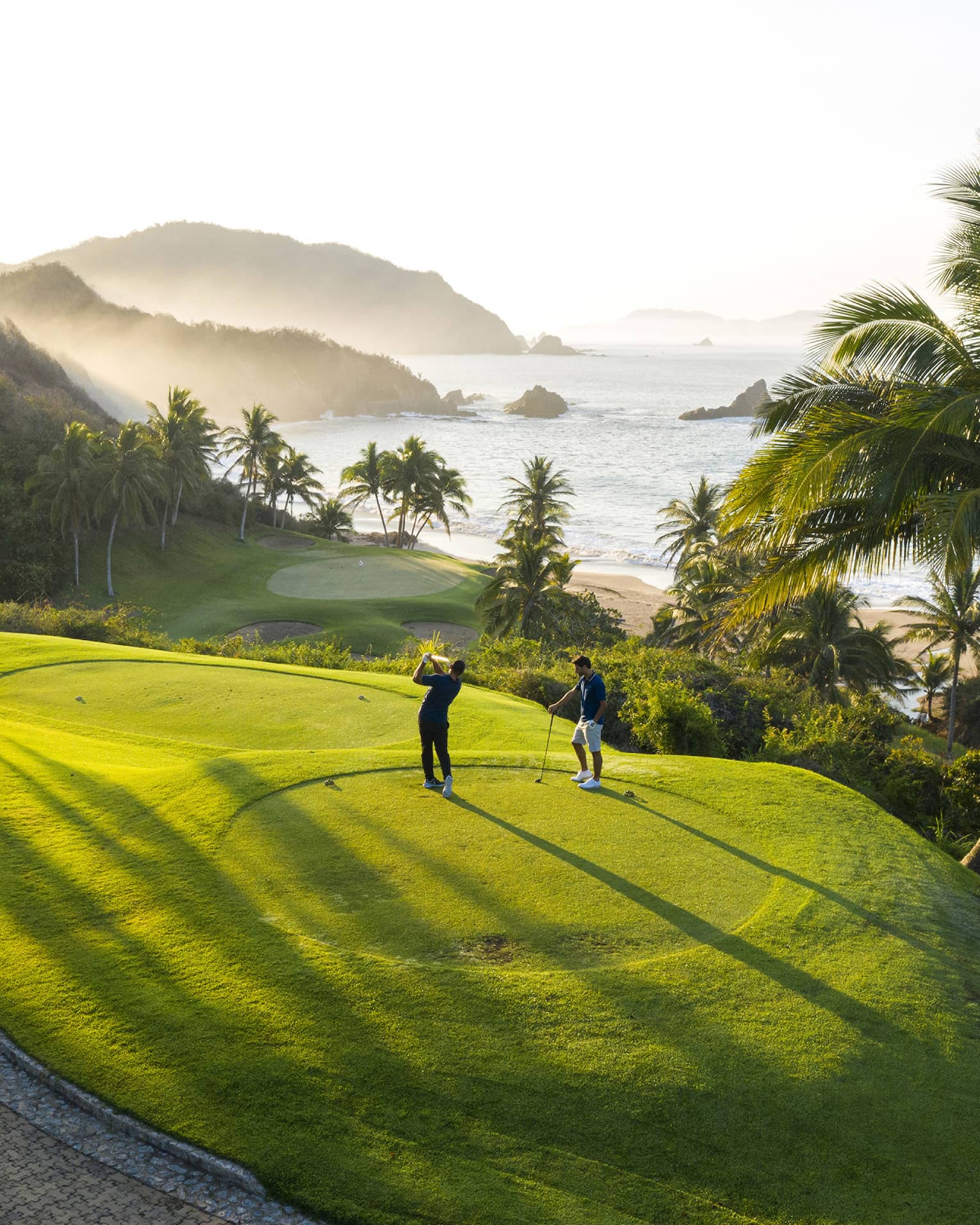 Rear view of two golfers on an elevated tee, one swinging toward the green down by the beach, misty hills and ocean beyond.
