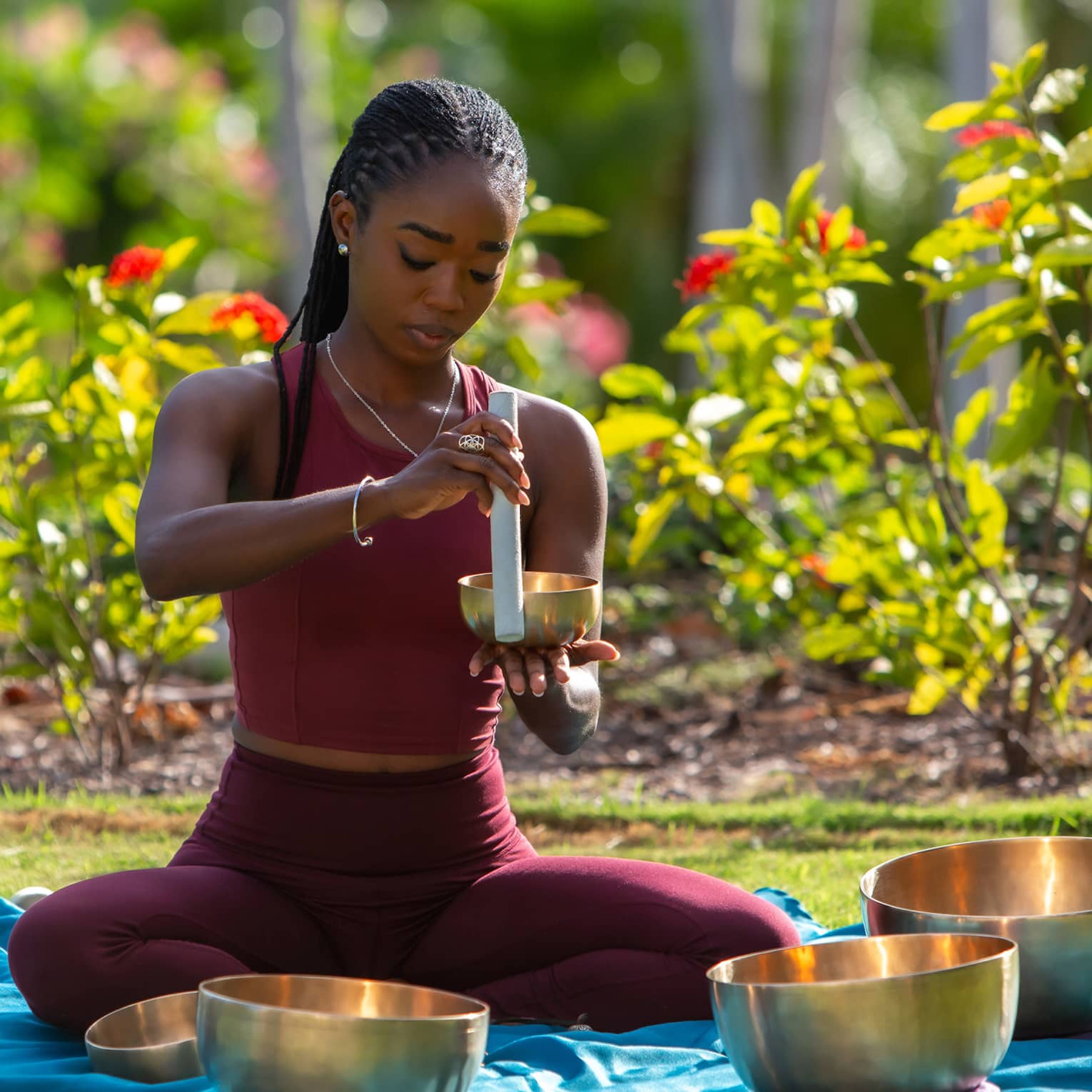 In a sunlit garden, a sound-bath practitioner sitting on a mat in a circle of brass bowls holds a tube against a small bowl.