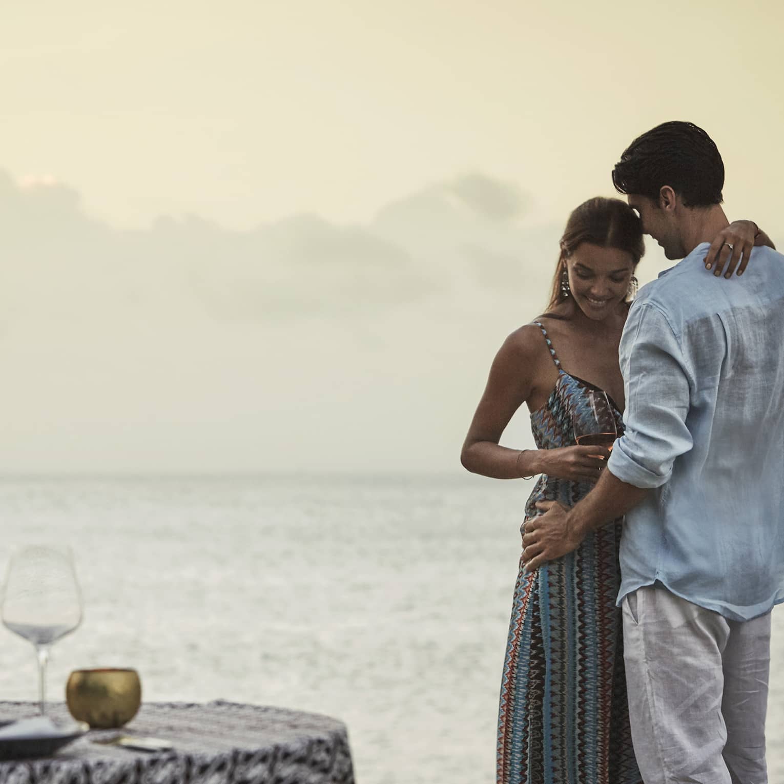 Couple embraces beside private dining table overlooking ocean at sunset