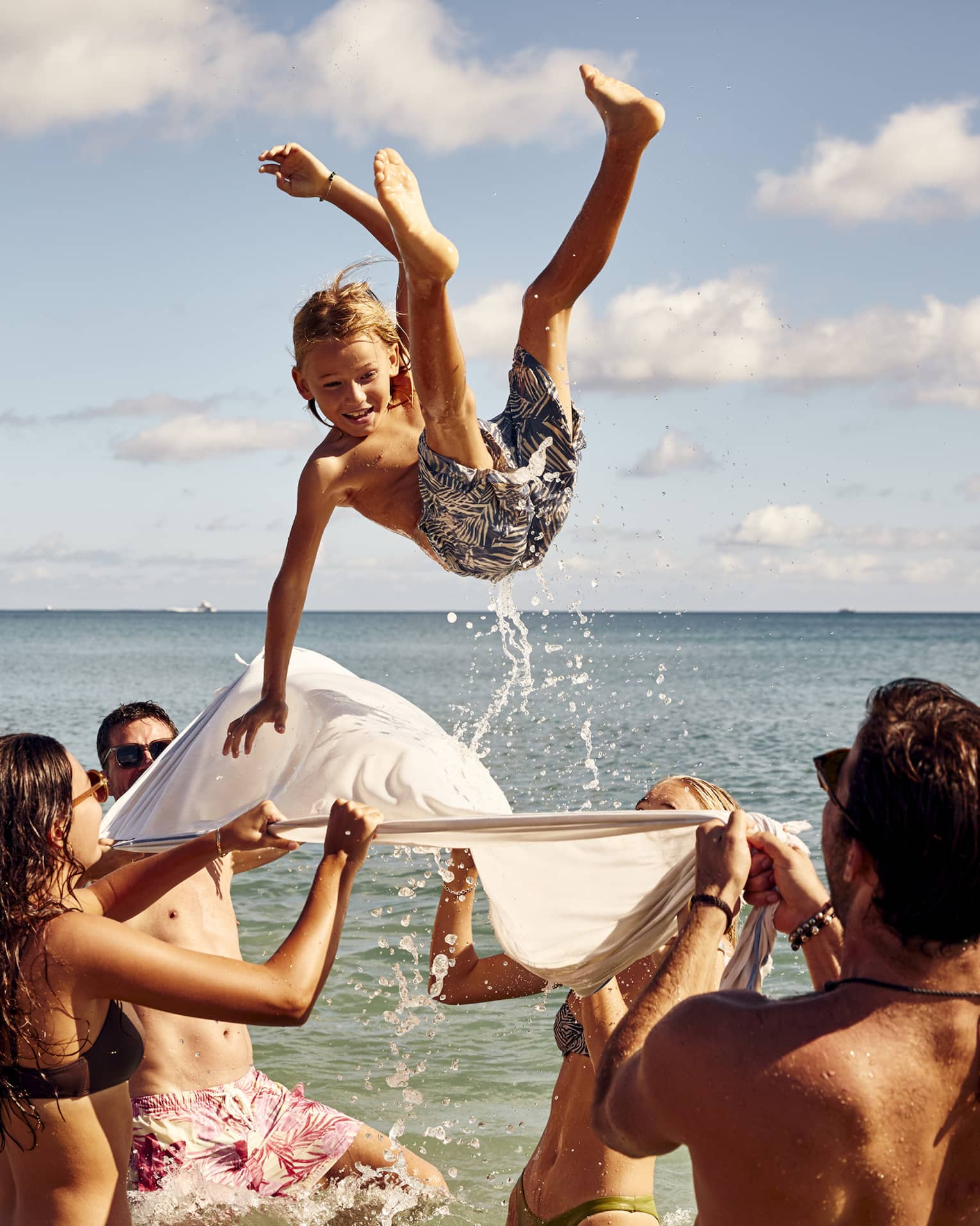 A group enjoys a sunny beach day, tossing a child into the air with a white sheet. The child smiles mid-air against clear skies.