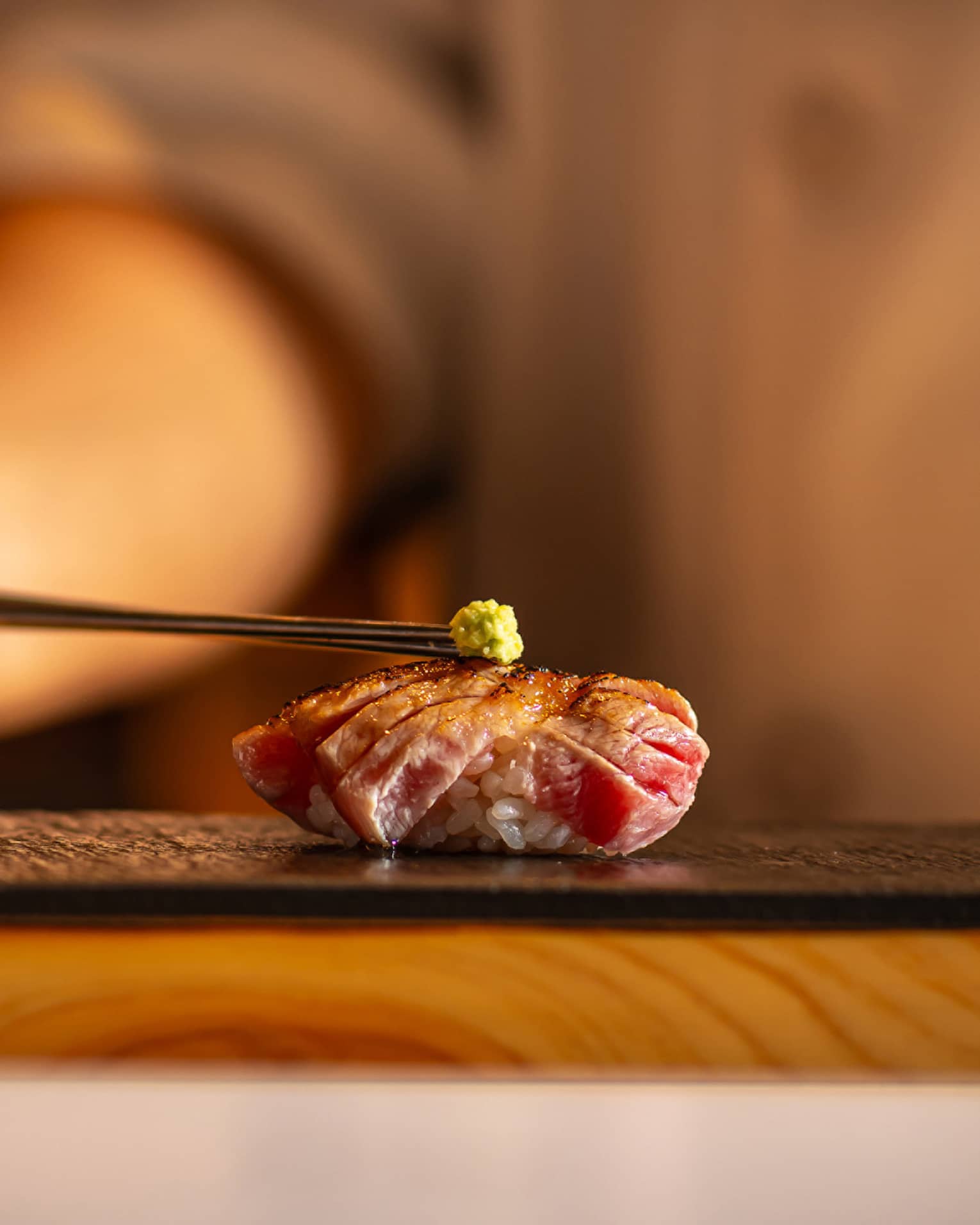 Seared tuna nigiri topped with a small dab of wasabi, placed on a black slate plate with a warm, blurred background