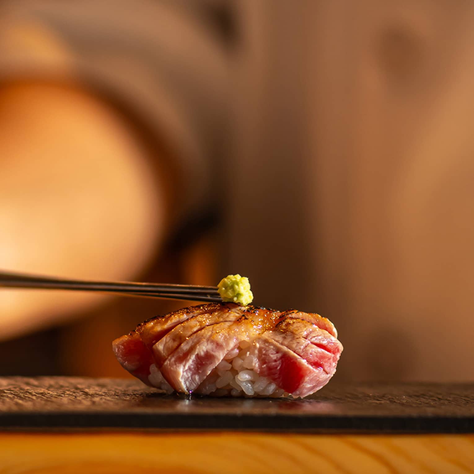 Seared tuna nigiri topped with a small dab of wasabi, placed on a black slate plate with a warm, blurred background