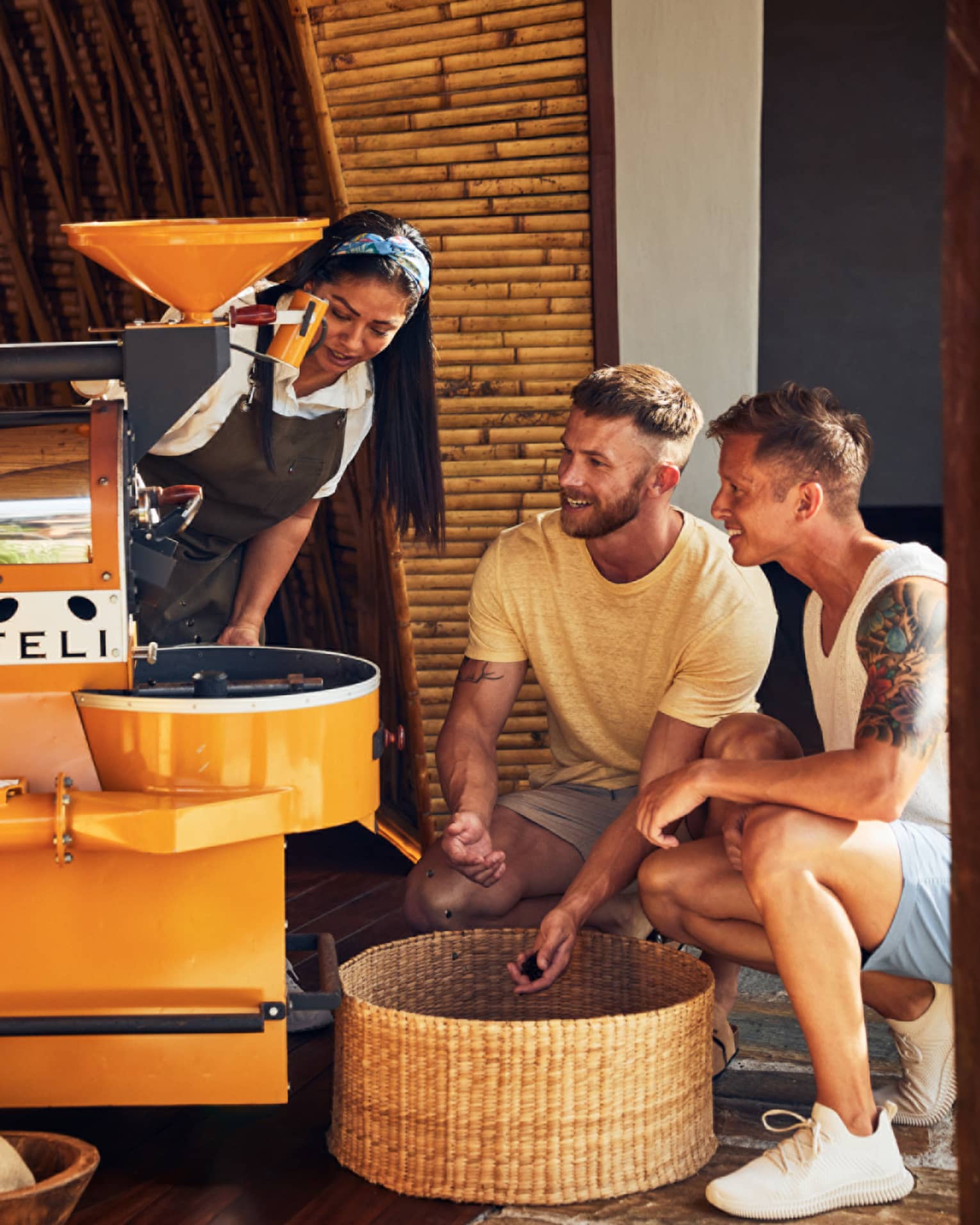 A woman demonstrates coffee roasting to two guests, who observe closely near a yellow coffee roaster in a bamboo-walled setting.