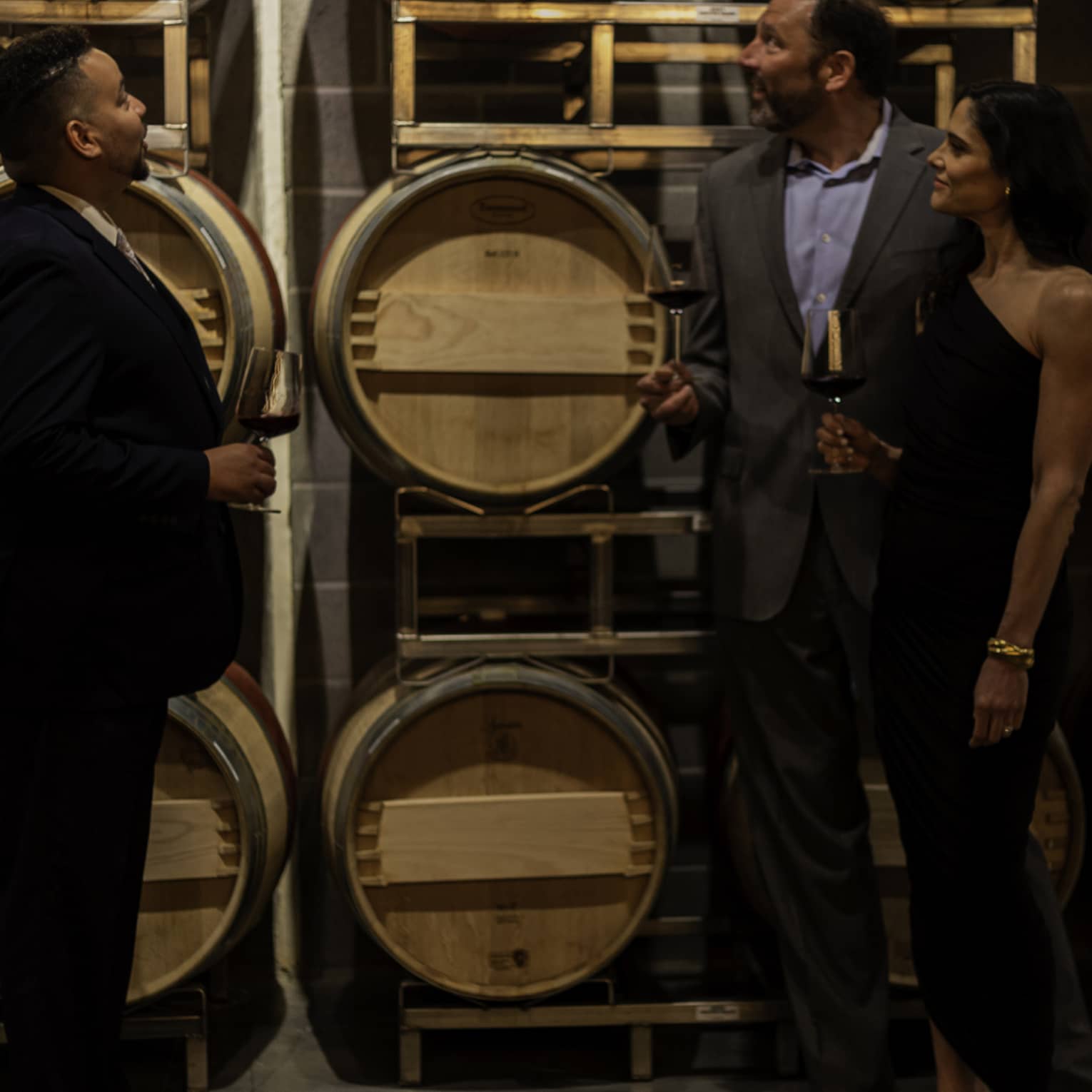 A couple in evening wear hold glasses of red wine and listen to a sommelier while looking at a rack of wine barrels.