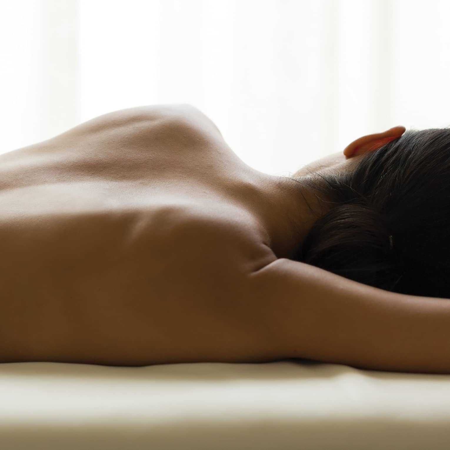 Woman's bare back as she lays on massage table under window, white curtains