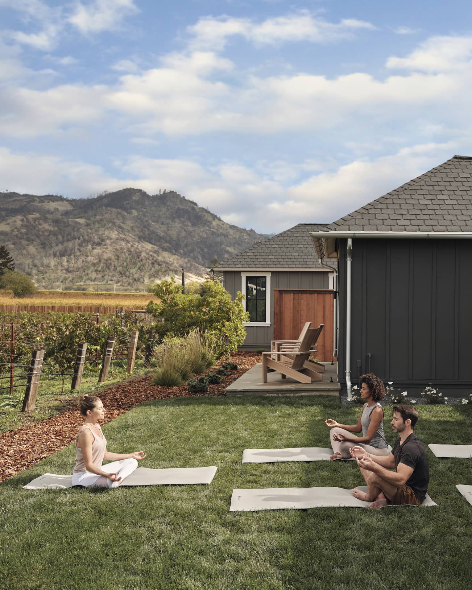 Group of people practicing yoga on mats in a grassy villa backyard near vineyards, with mountain views, wooden chairs and an outdoor grill area under a pergola