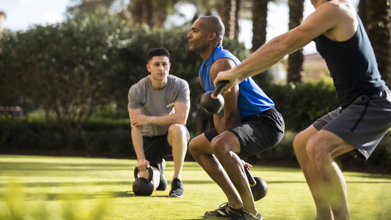 Four Seasons trainer watches as two men squat, lift kettlebell weights on lawn
