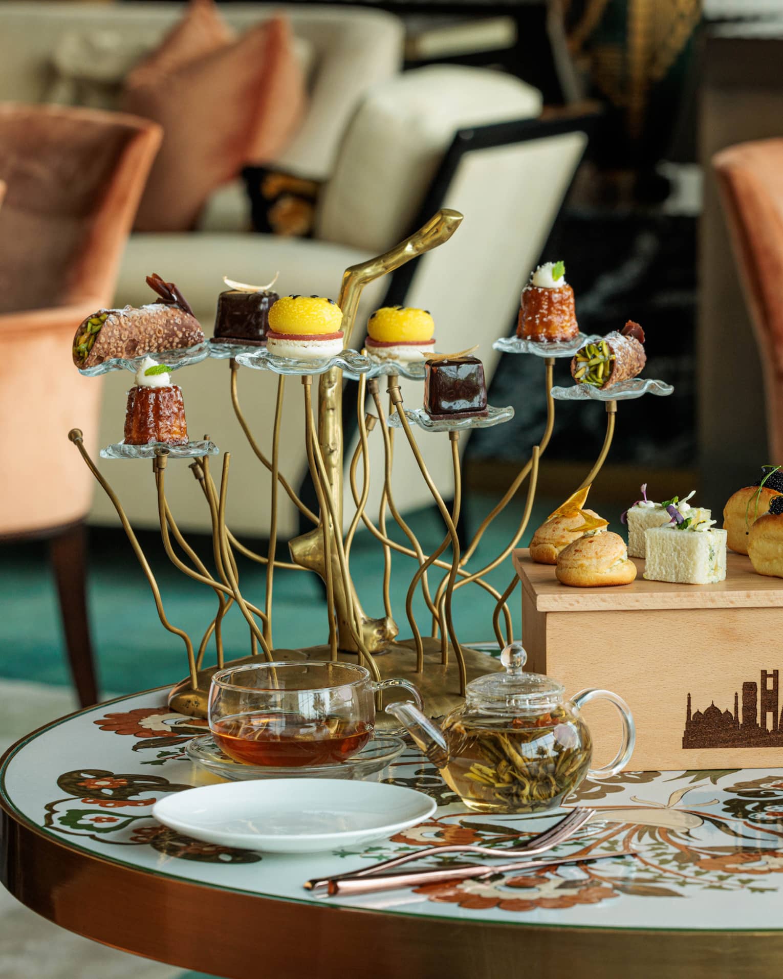 Table set for afternoon tea featuring a glass teapot and cups, various pastries on a wooden box and a tree-like pastry stand with a pastry on each branch