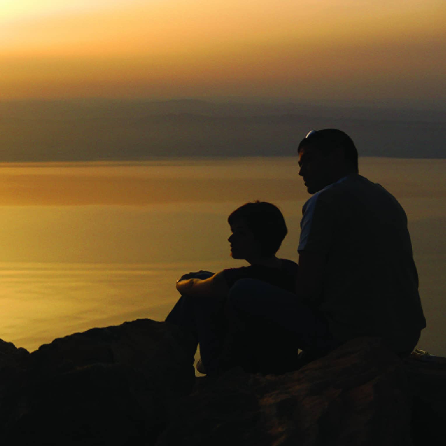 A couple sitting on a rocky outcrop are silhouetted against a setting sun that casts an orange glow on the lake before them.