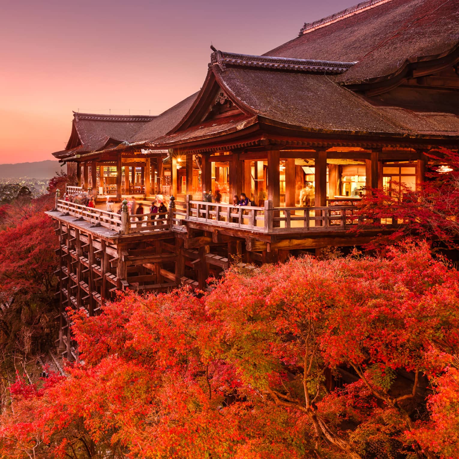 Massive temple on stilts under a pink-hued sky, people on patio admire red-leafed trees, distant city skyline and mountains.