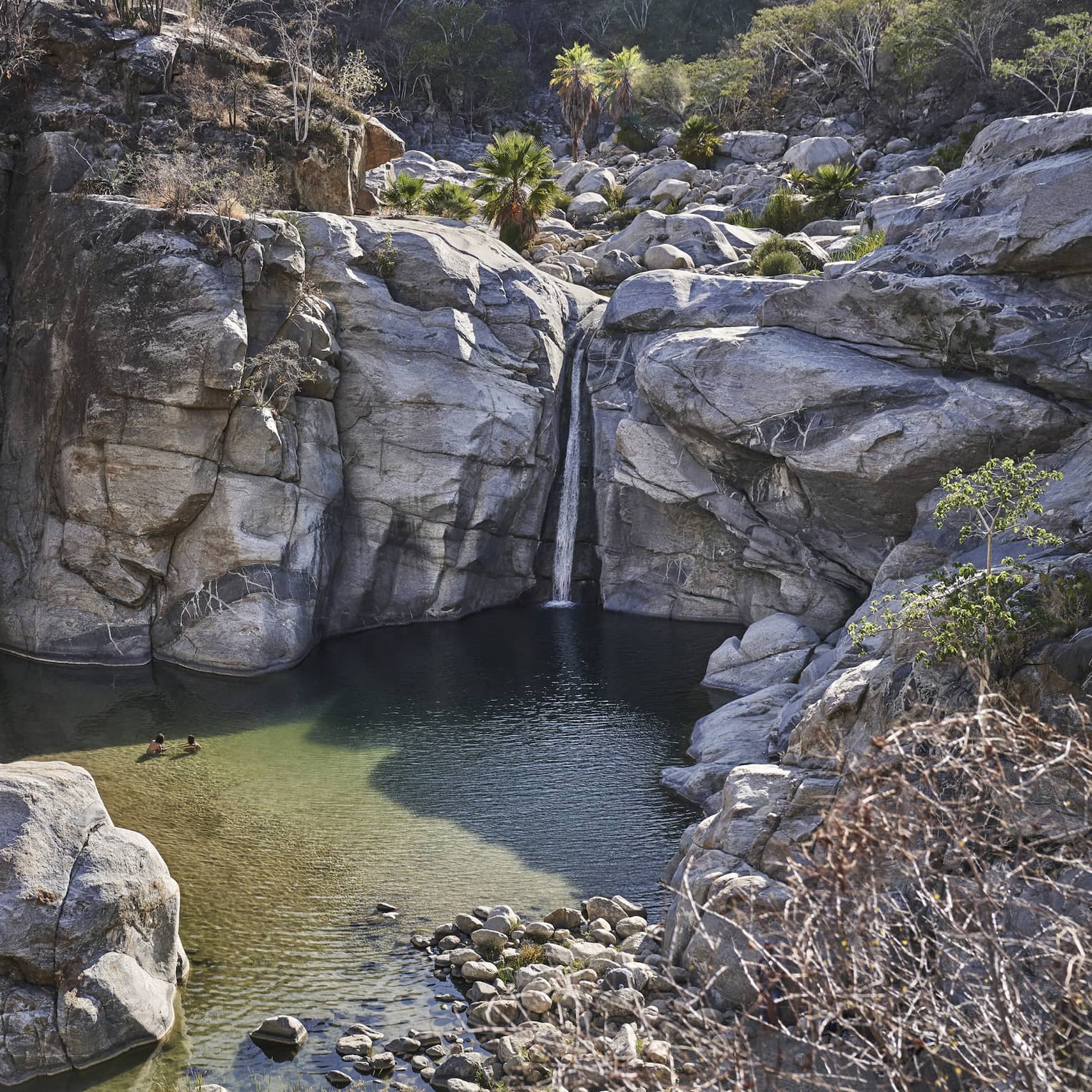 Two people wade in a small ocean lagoon surrounded by large cliffs, creating a serene and picturesque scene with clear water and rugged natural beauty.