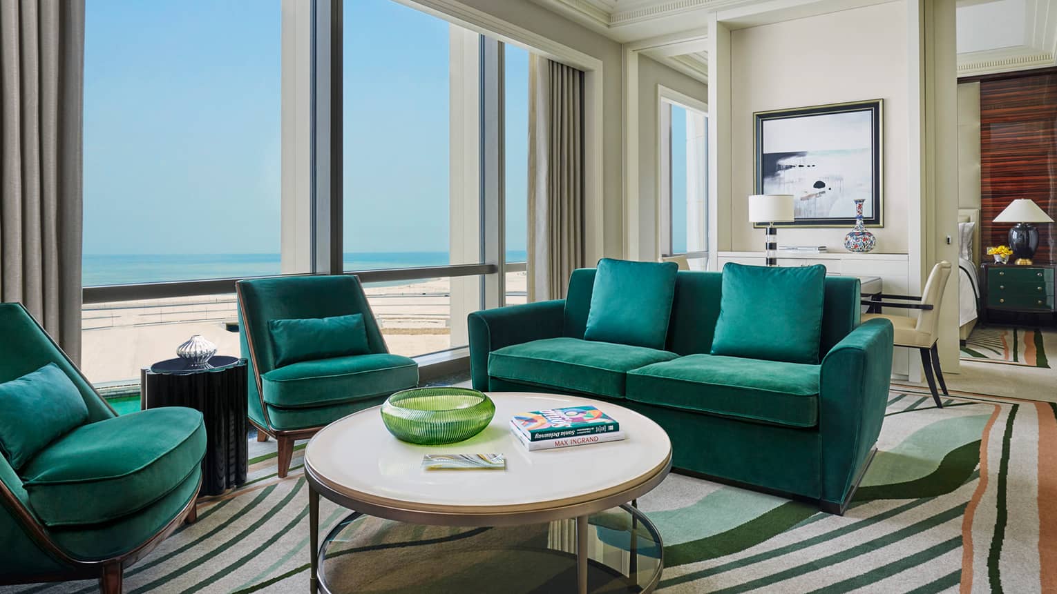 Four Seasons Executive Suite with emerald green velvet sofas, armchairs, round coffee table with books, sunny windows