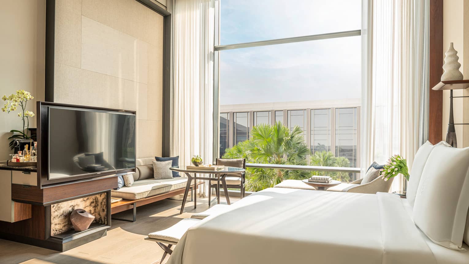Hotel room with white bed facing a TV console, off-white wall and wall of windows, high ceilings