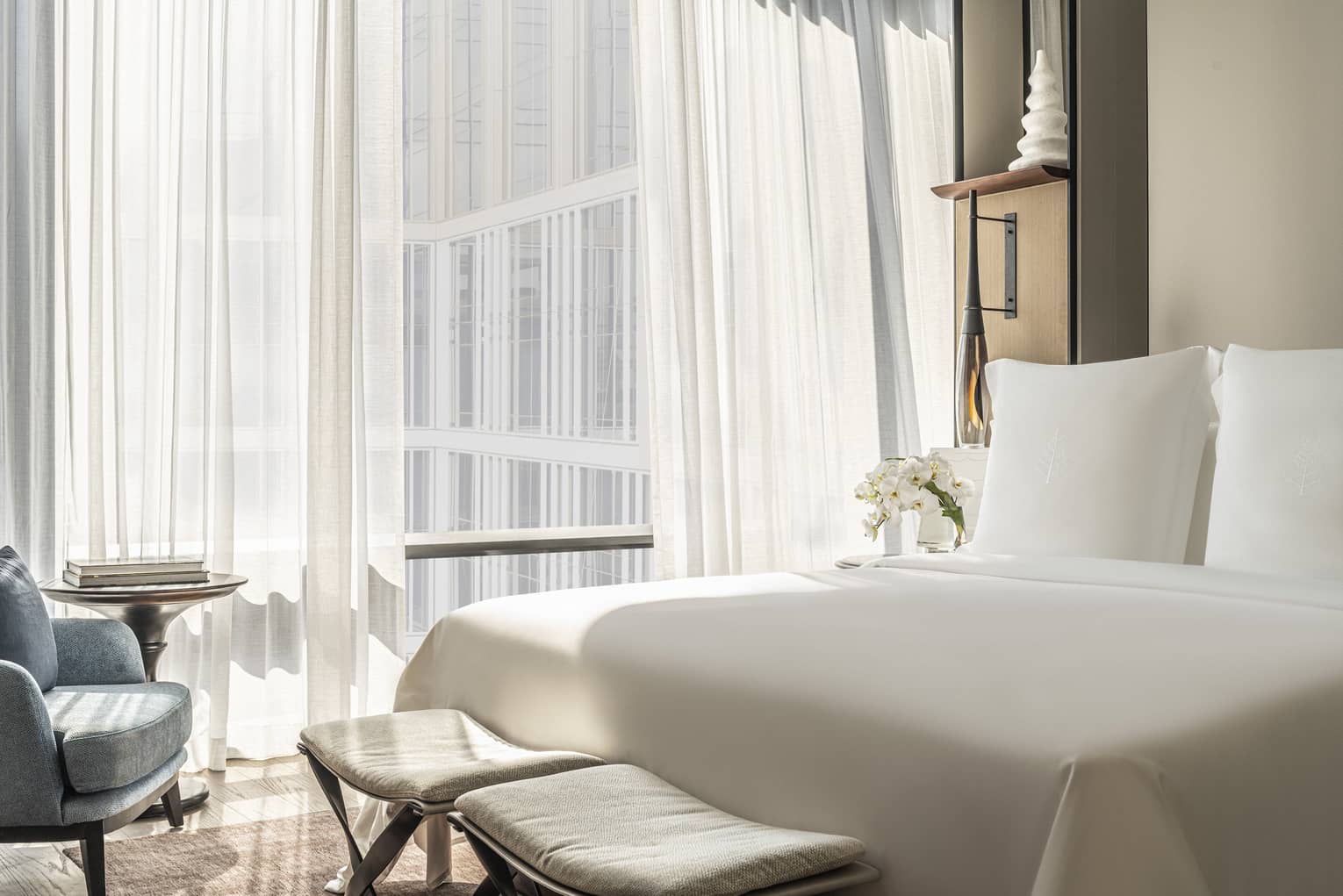 Hotel room with white bed and two benches at foot, facing blue chair, off-white wall by curtained window