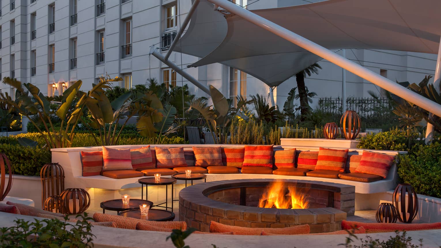 Large curved white bench with cushions around outdoor fire pit with flames at dusk