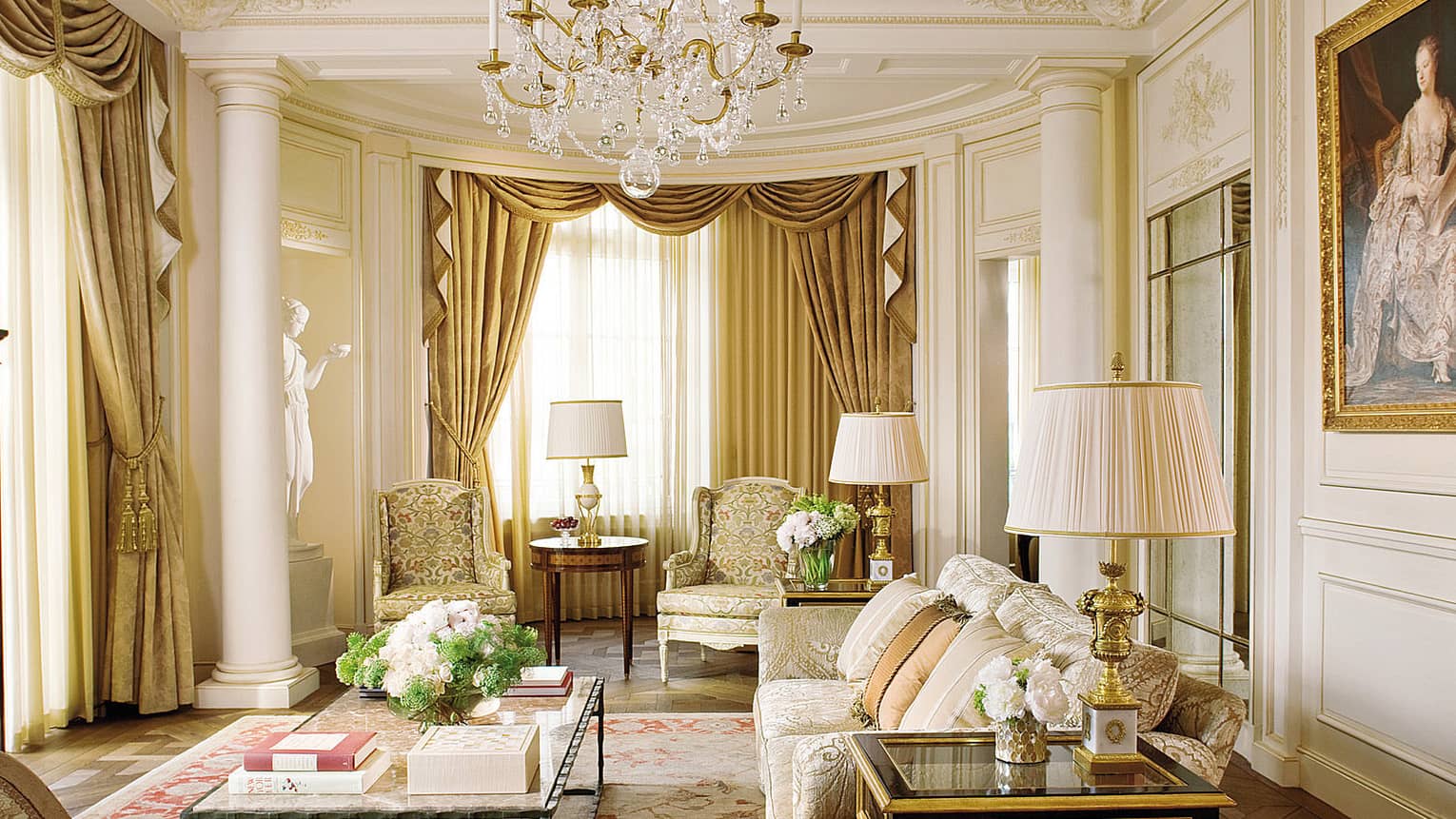Luxury sitting room with gold accents, ornate drapery, armchairs, chandelier and floral centrepiece on a coffee table, framed by grand columns and a statue
