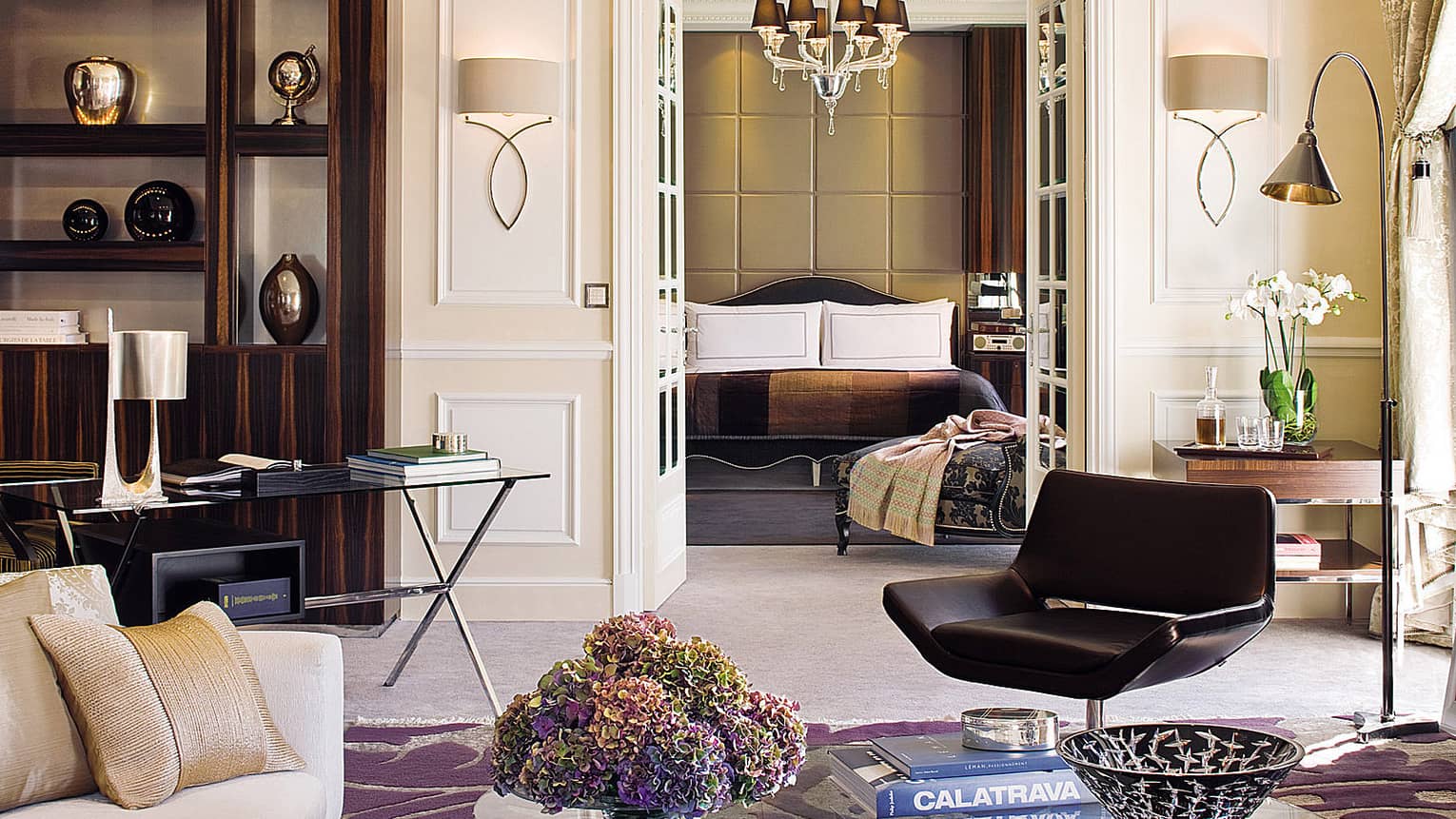 Hotel living room with coffee table, book, retro black chair, glass desk in front of shelf, vases, bedroom door