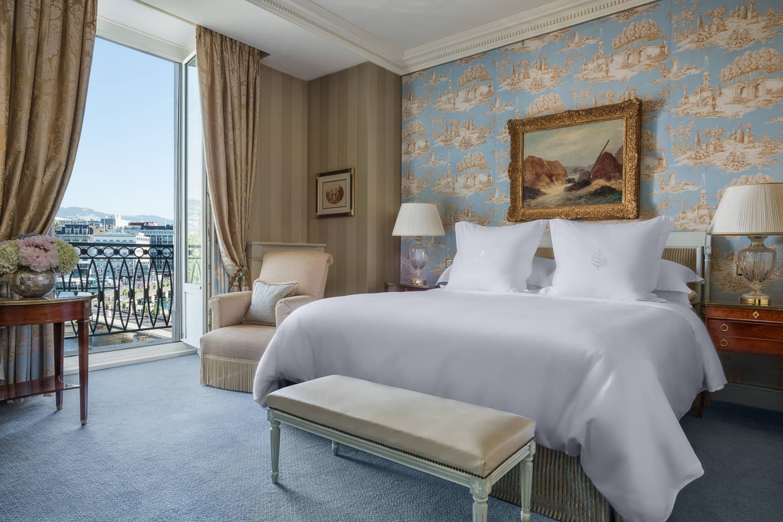 Luxury hotel room with a king bed, nightstands with lamps, bench, patterned wallpaper, armchair, framed artwork and a balcony with city view