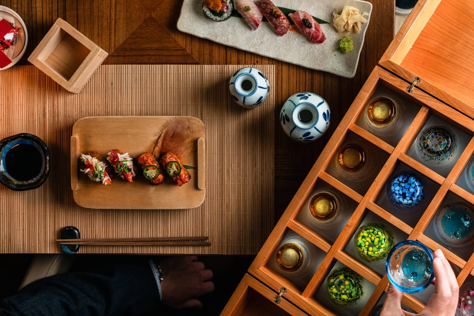 Aerial view of various nigiri and sushi, a bamboo placemat, chopsticks, and soy sauce served in a blue ceramic dish, next to a wooden box filled with various colourful sake glasses