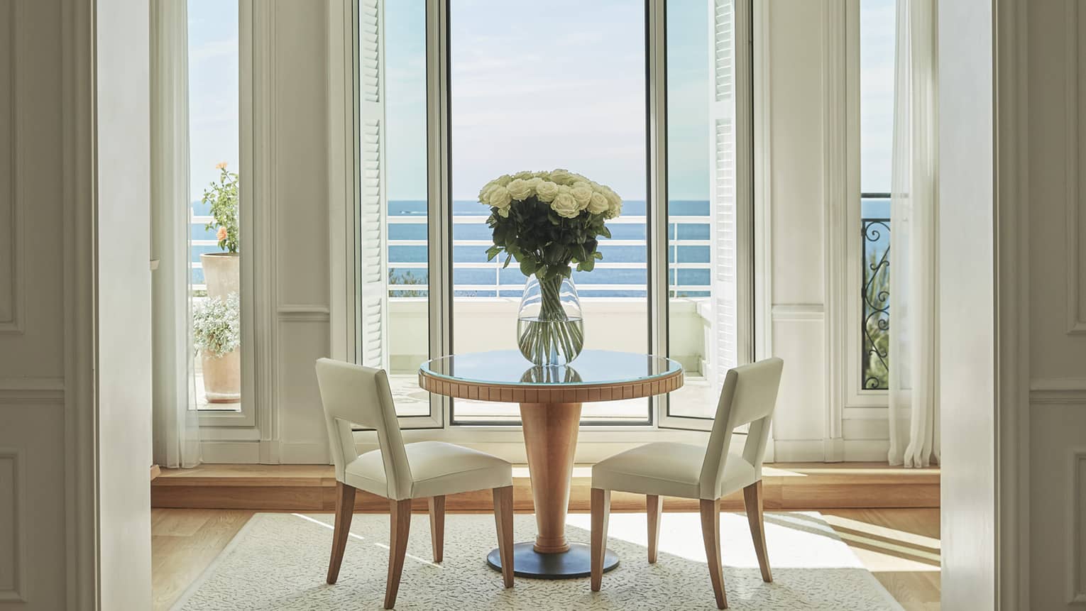 White chairs by small cafe table, large glass vase with flowers, sunny window