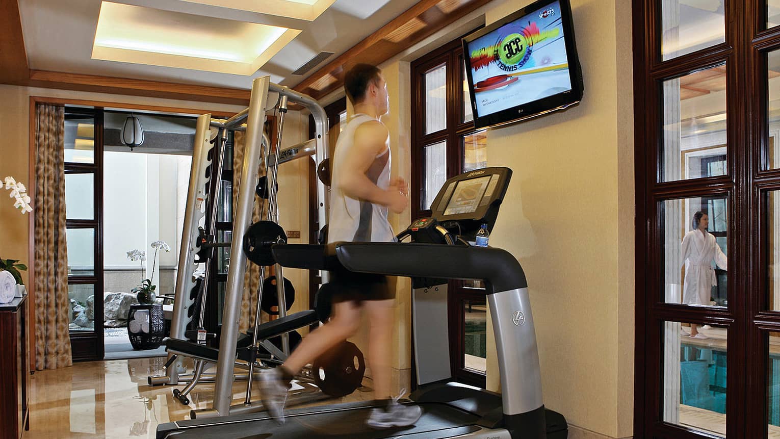 Man running on treadmill in Fitness Centre, woman in robe walking by pool in background