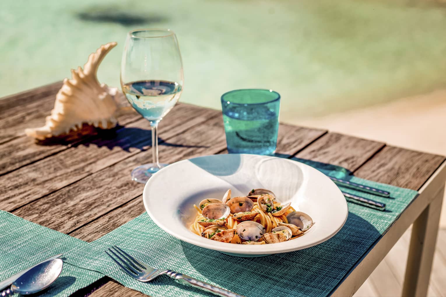 Spaghetti alle vongole on wooden picnic table with turquoise placemat and water glass, clear wine glass, conch shell