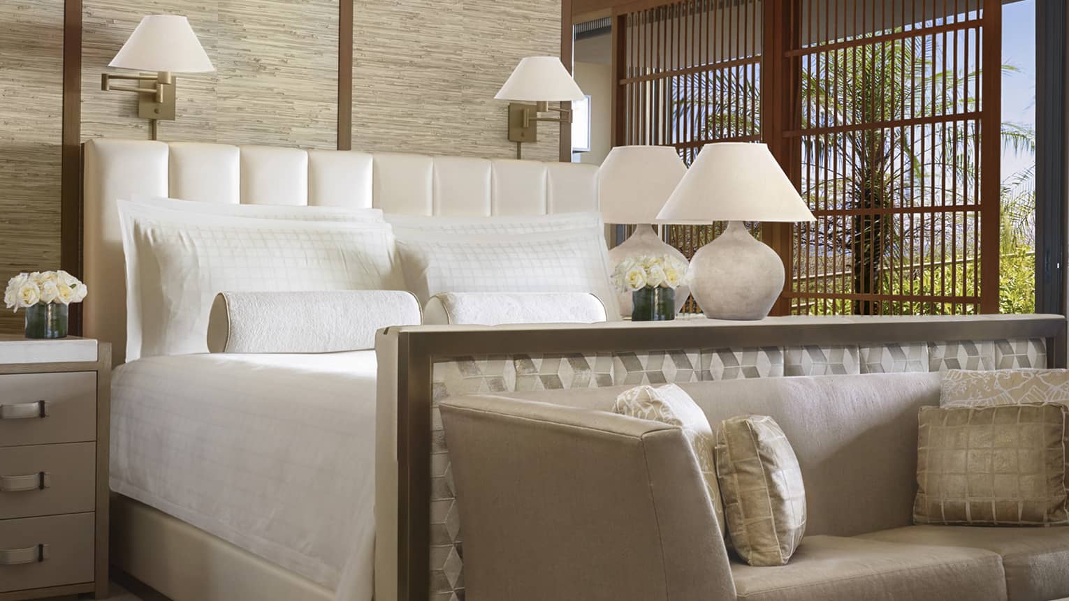 Makai suite close-up view of bed with white linens, fabric footboard with small grey loveseat, sliding balcony door with wood shutter