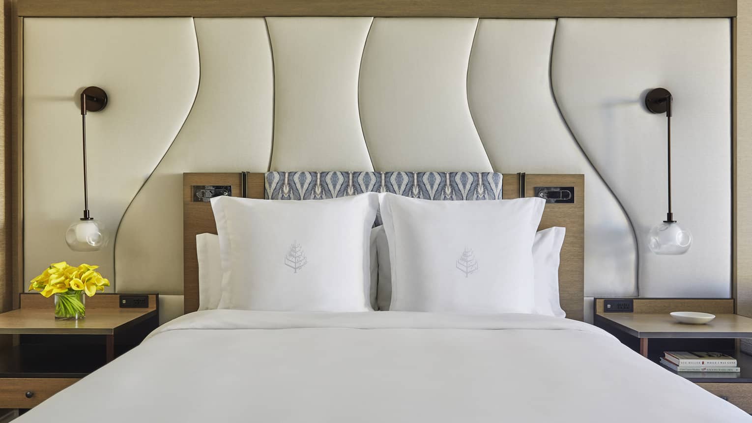 Modern bed with white linens, padded headboard, pendant lights and nightstands with flowers and decor