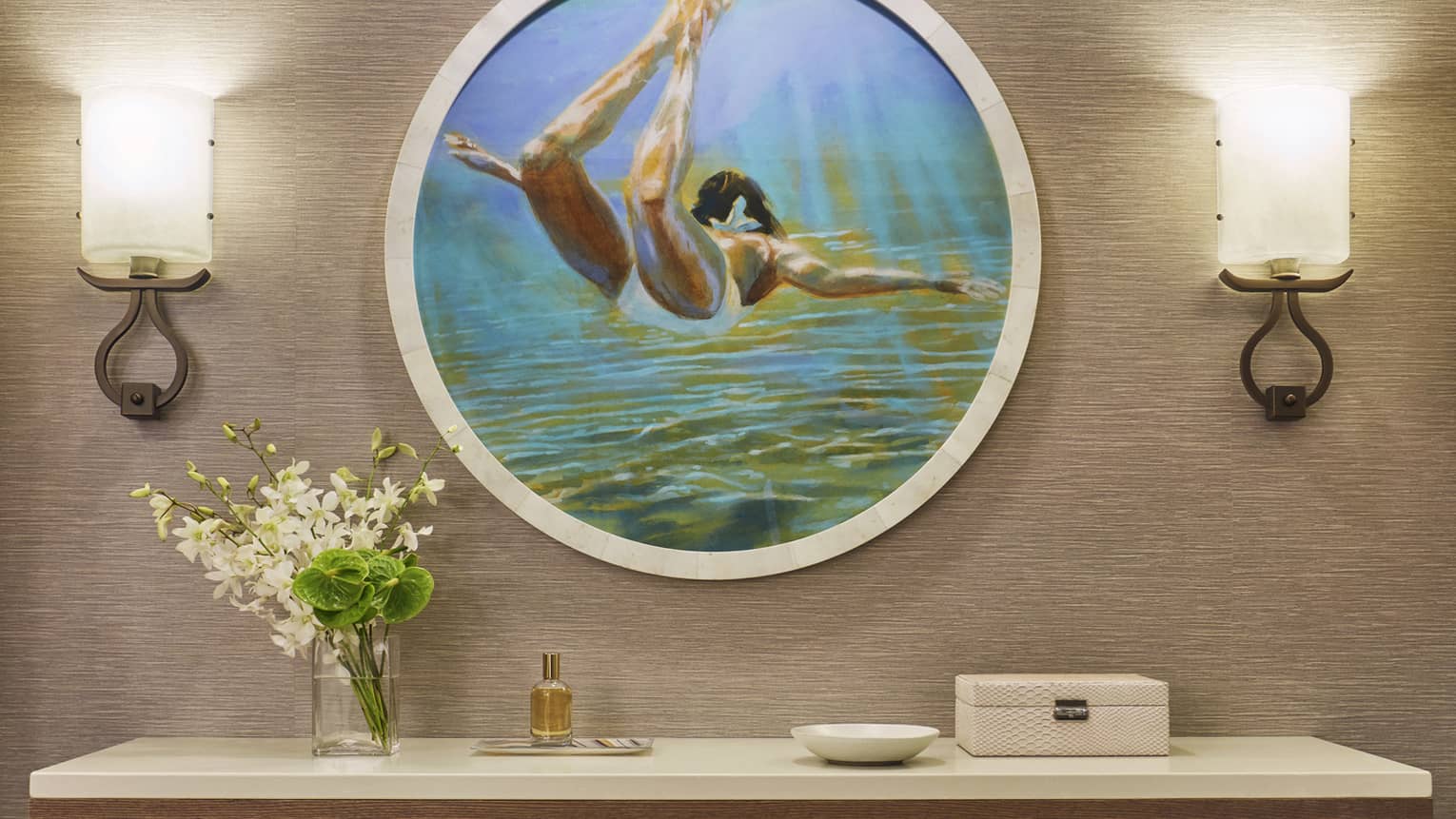 Wall-mounted console with flowers, perfume and decor beneath round artwork of a swimmer and two wall sconces