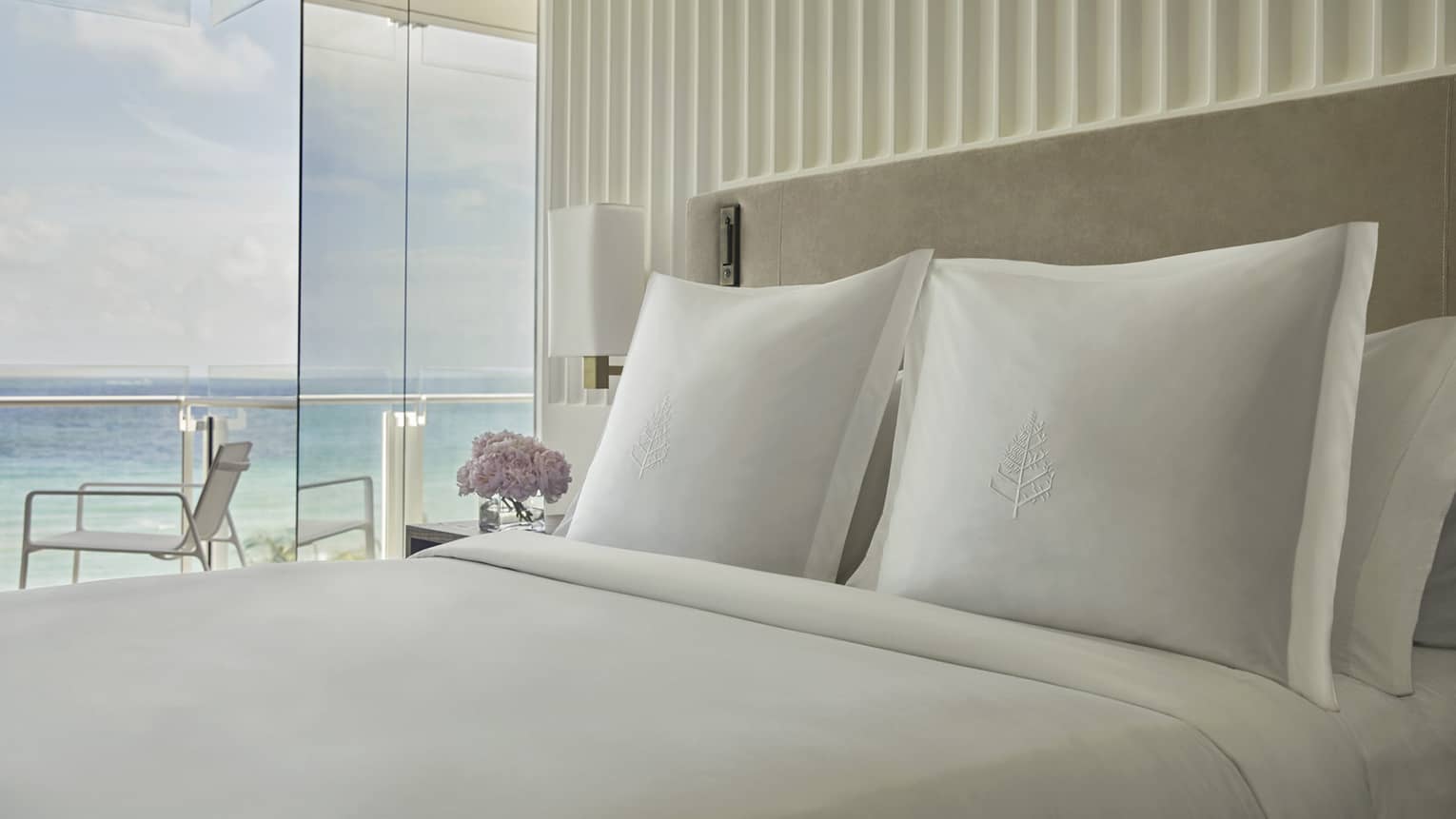 Luxury hotel bed with white embroidered pillows, a bedside table with flowers and floor-to-ceiling windows offering ocean views