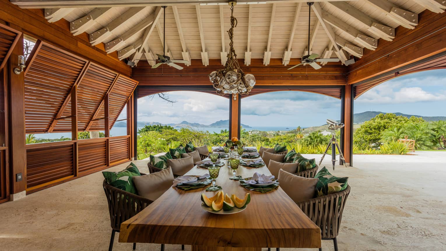 Melon slices on long rustic wood dining table, chairs and pillows under covered beach patio