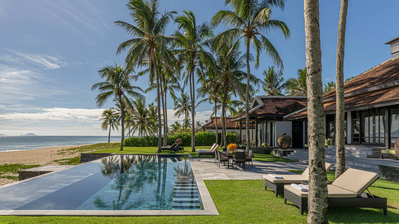 Three Bedroom Beachfront Pool Villa pool area with dining table, lounge chairs, palm trees and blue skies