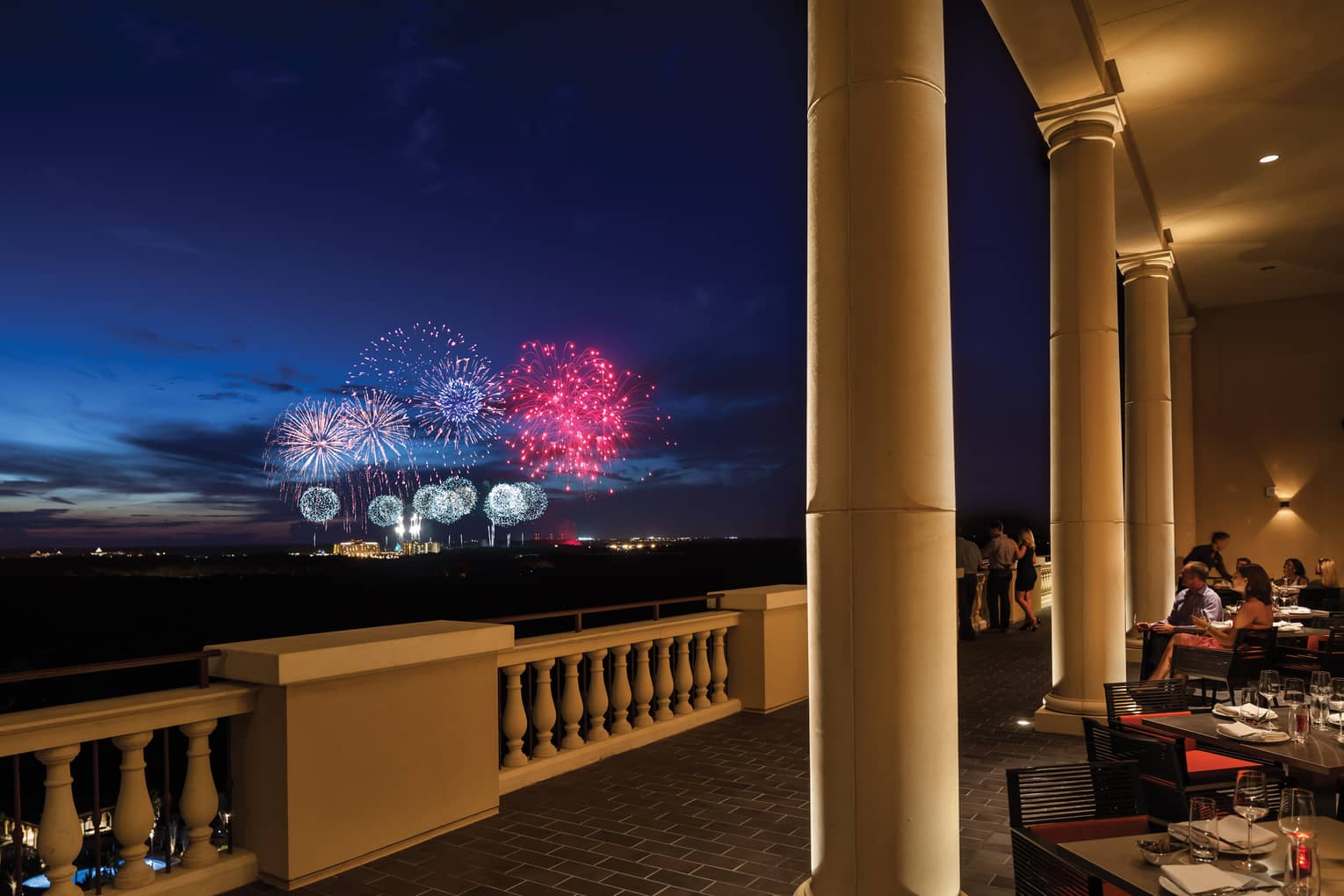 The terrace of a restaurant with tables and people watching fireworks in the distance.