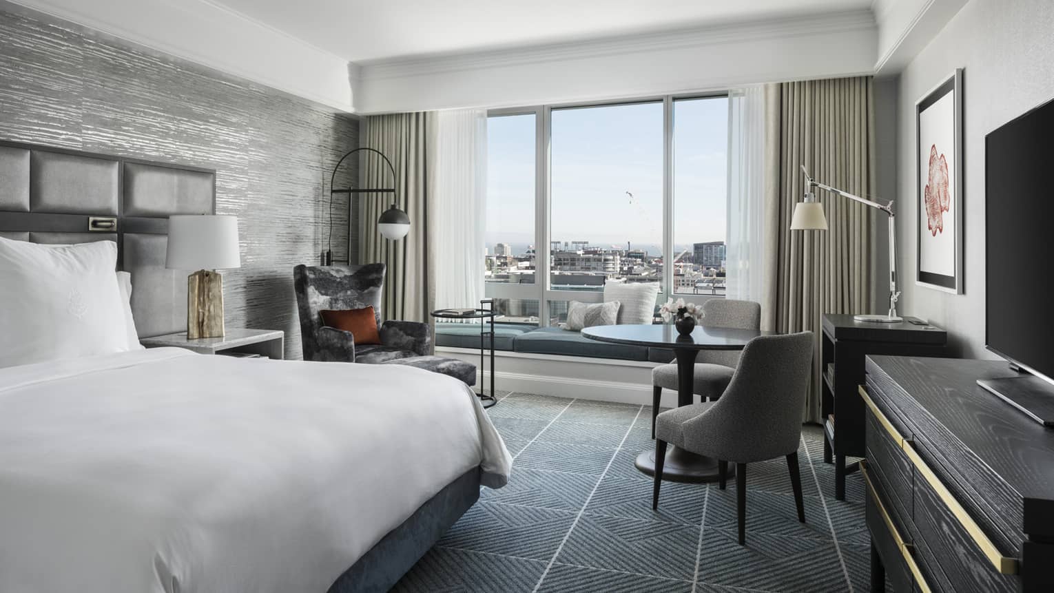 Modern hotel room with king bed, table and two upholstered grey chairs, windows overlooking San Francisco